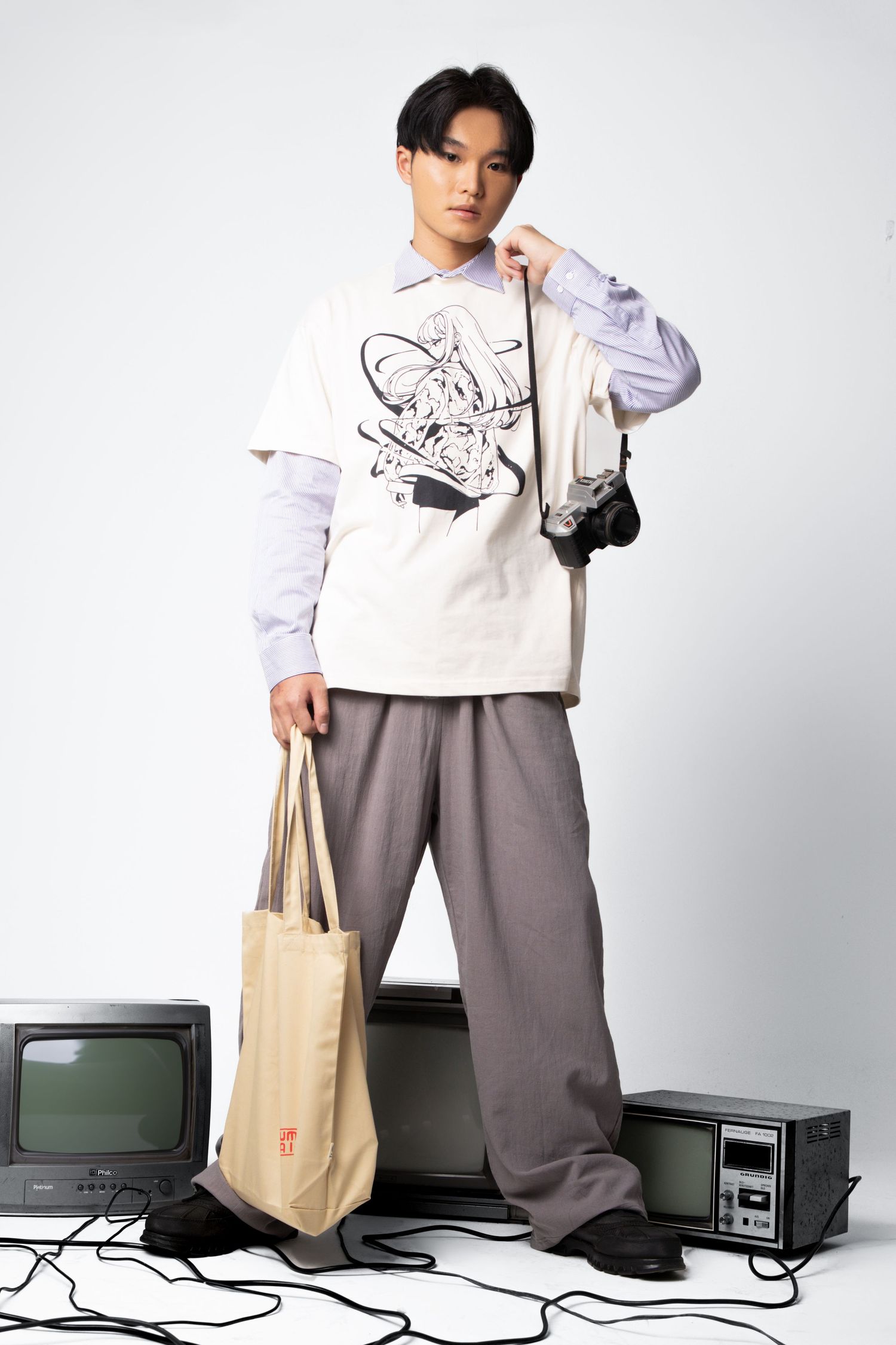 A young man in a graphic cream tee, layered shirt and grey trousers holds a tote bag and camera beside vintage televisions.
