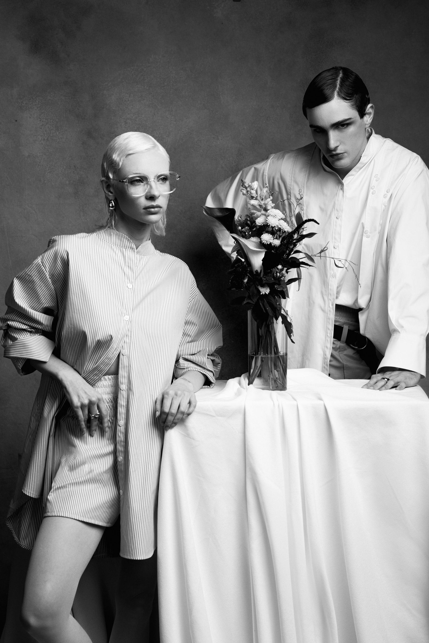 Black and white editorial of two models in loose white shirts beside a draped table holding a floral arrangement.