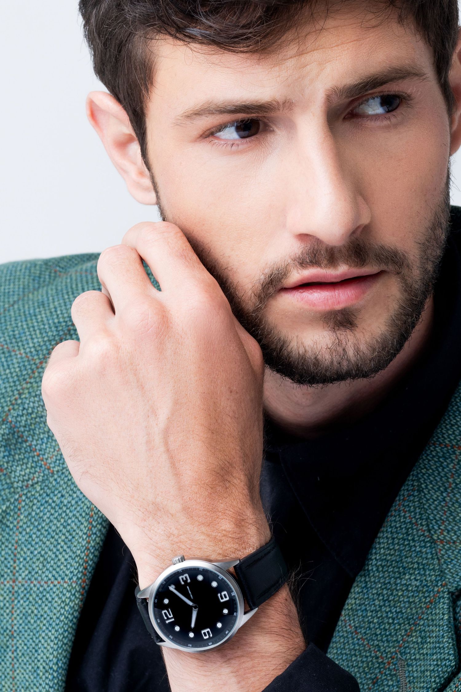 Close-up of a bearded man in a green tweed blazer touching his face, wearing a black dial wristwatch with a black leather strap.