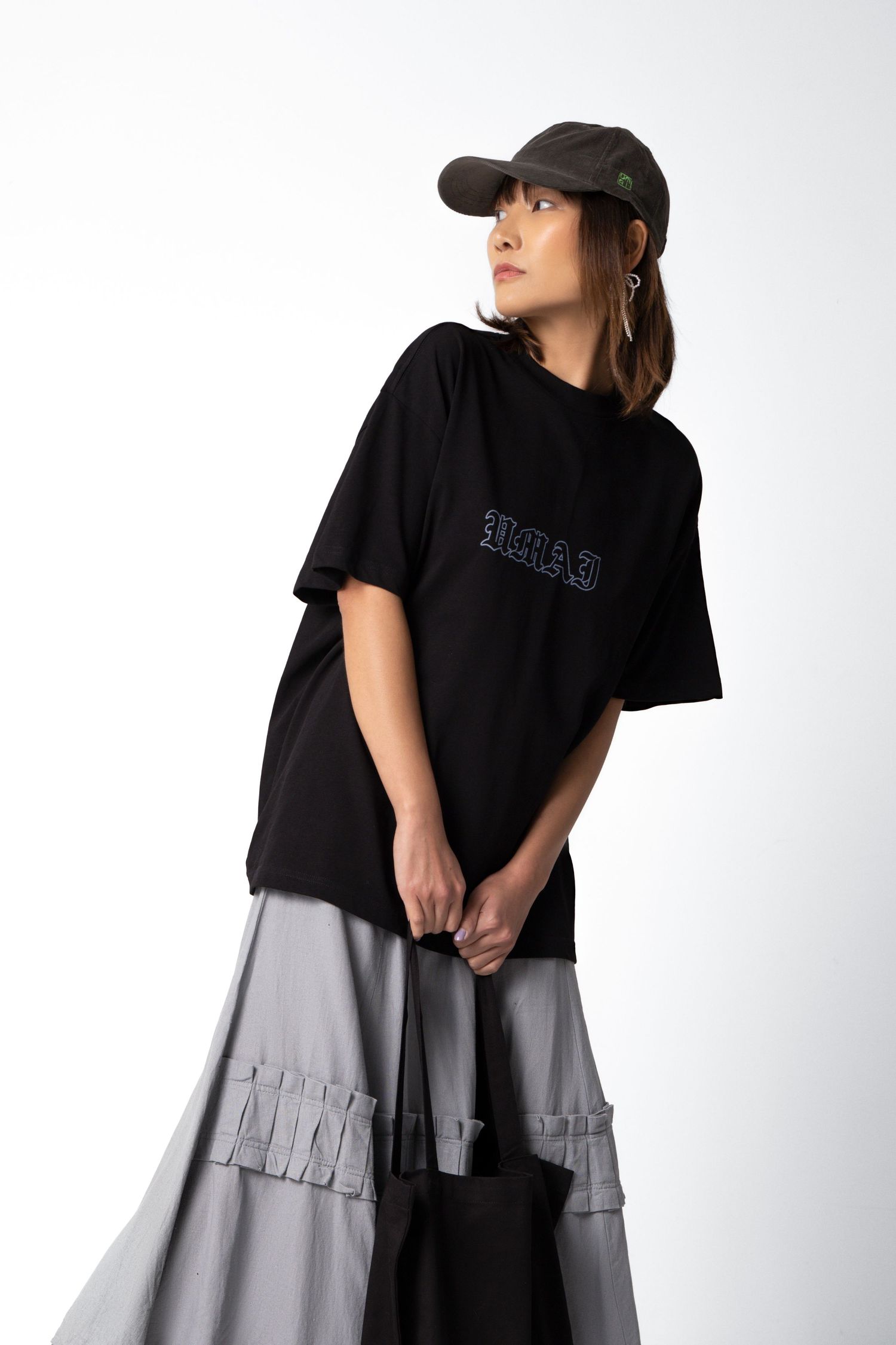 Close-up of a woman in a black oversized graphic tee, gray ruffled skirt and dark cap holding a black tote bag.