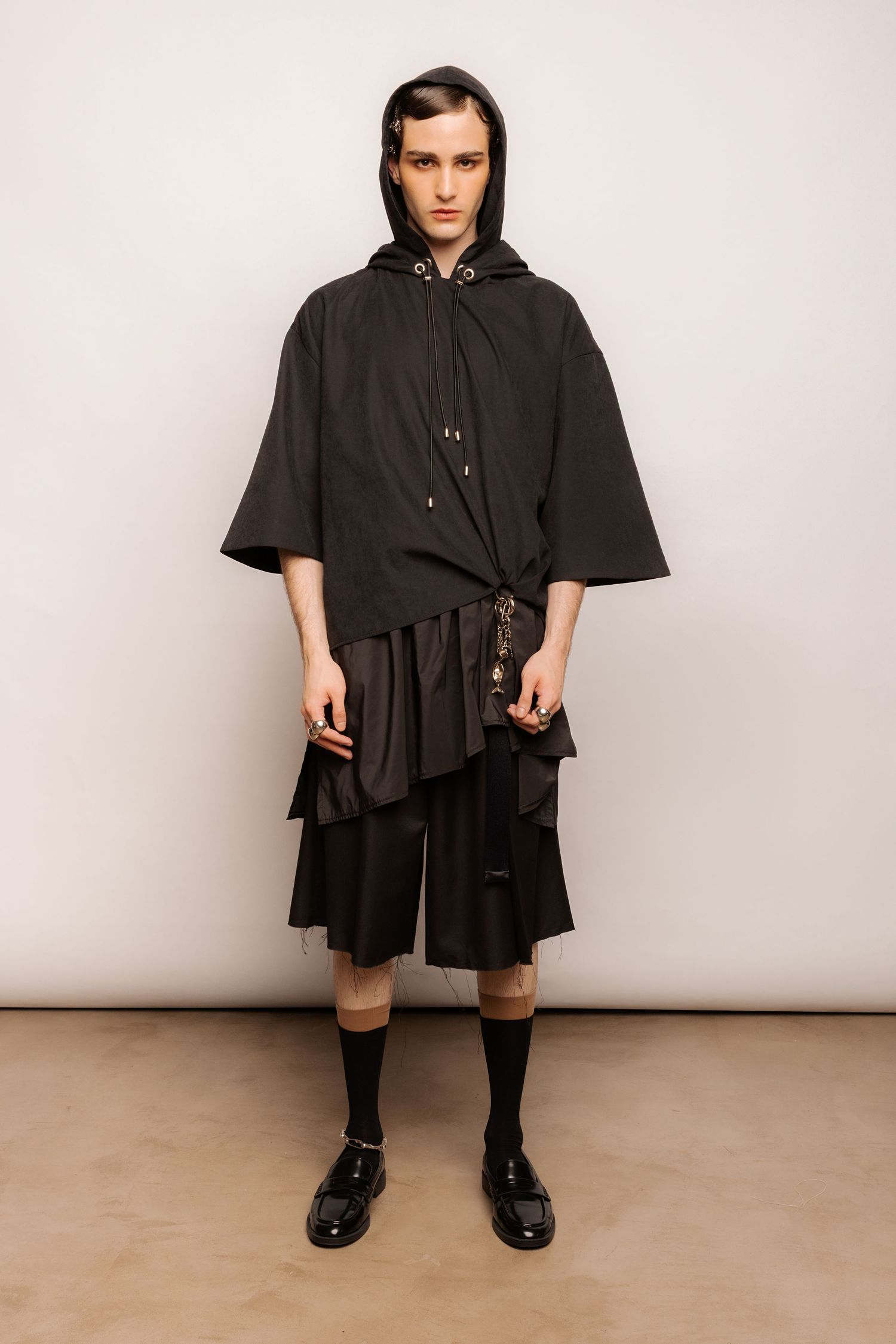 Model in a black hooded cape-style top, layered pleated shorts, tall black socks and black loafers posing in a studio.