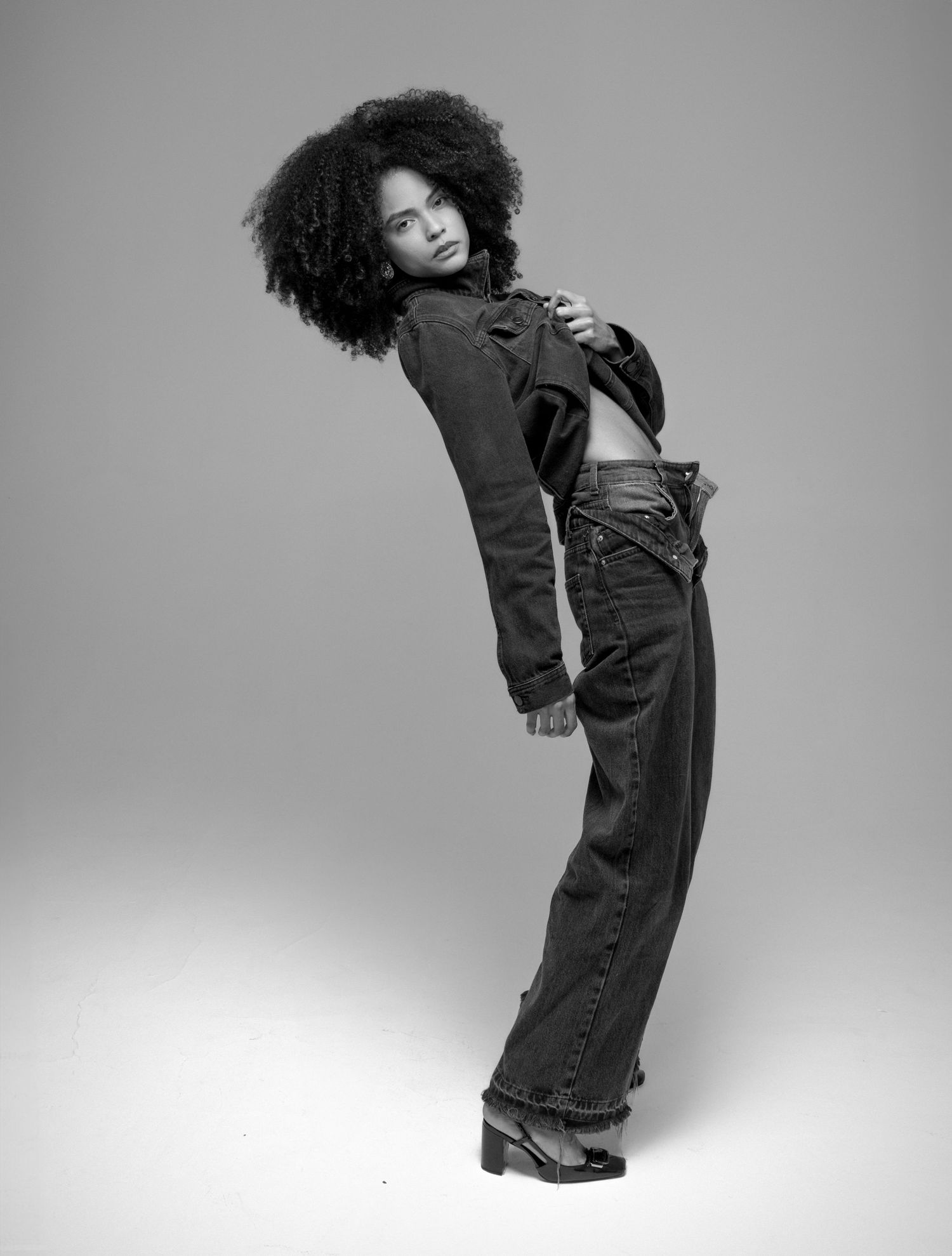 Black and white image of a model with voluminous natural curls in a denim jacket and wide-leg jeans leaning back in a dynamic pose.