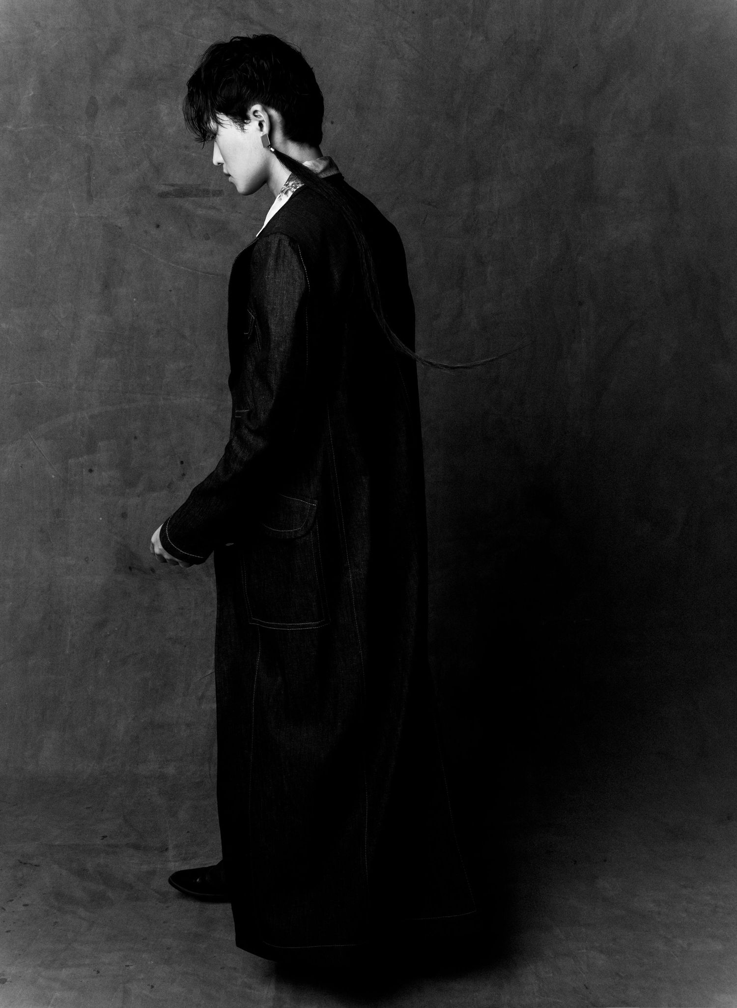 Black and white portrait of a model in a long dark coat standing in profile with head turned down against a textured studio backdrop.