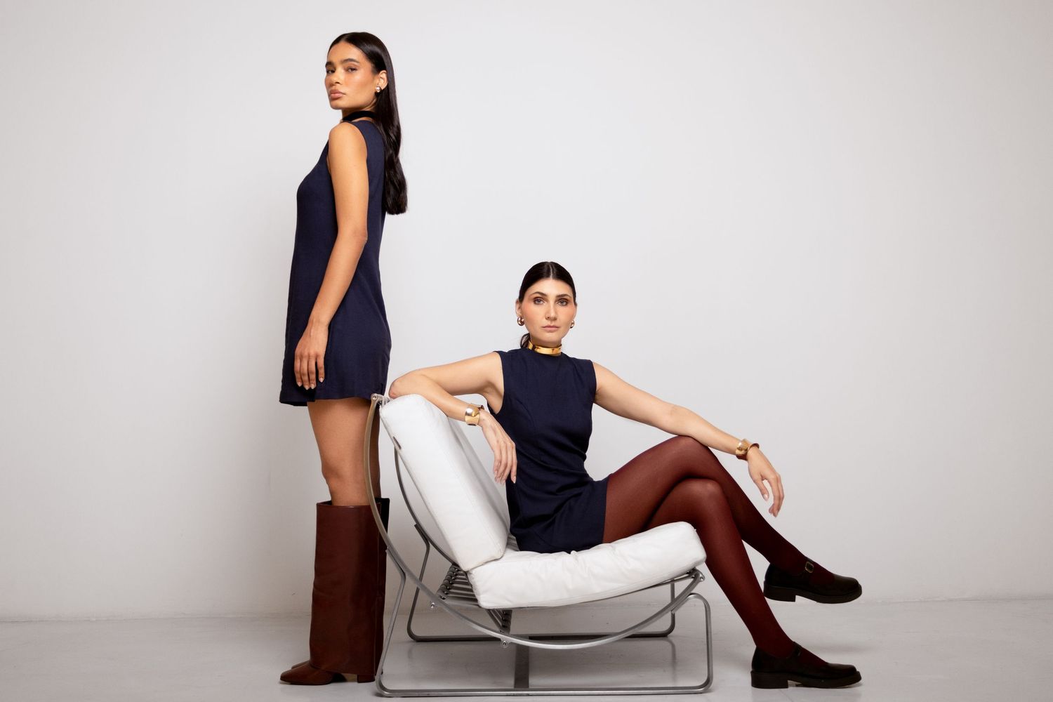 Two women in navy dresses pose in a white studio, one standing in tall burgundy boots, the other seated in a chrome and white chair.