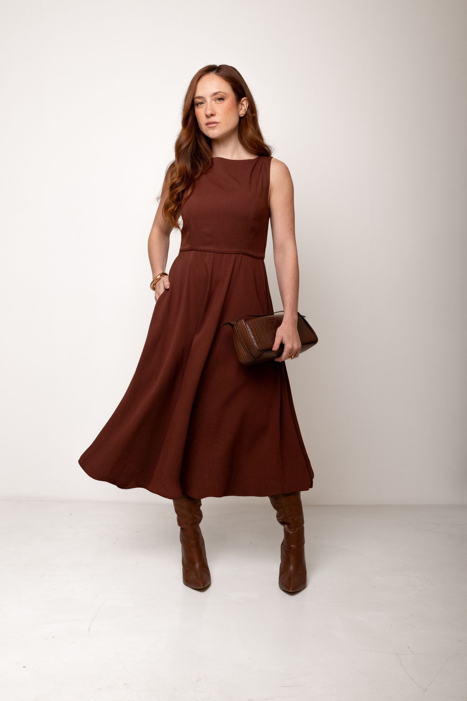 Woman in a rust brown sleeveless midi dress with a flared skirt, styled with a brown clutch and tall brown leather boots.