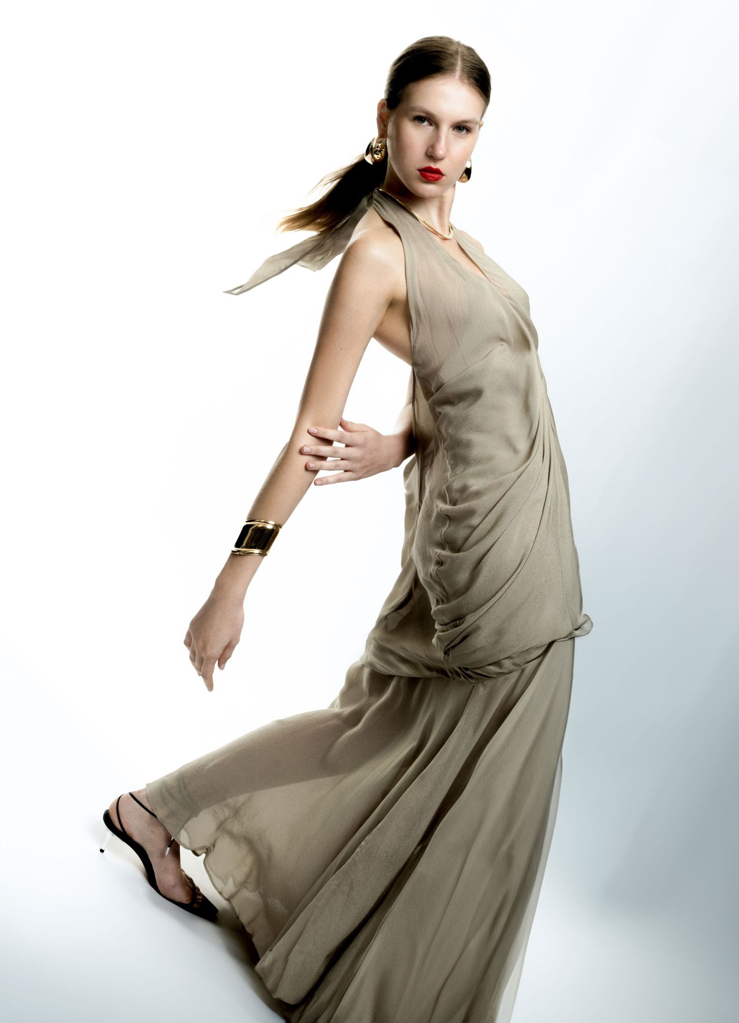 Model in a flowing olive halter gown with a ponytail, red lipstick and a gold cuff bracelet posing against a soft white studio background.