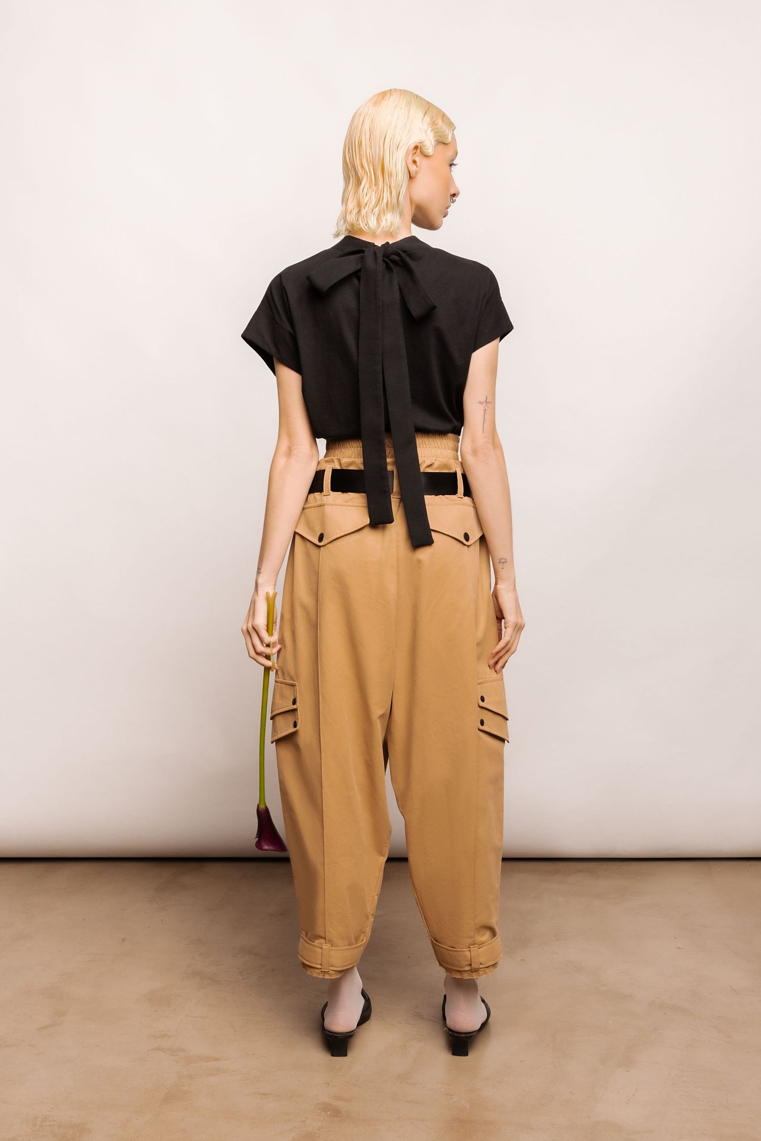 Back view of a model with blonde hair wearing a black bow-tie blouse, tan cargo trousers and black-and-white heels, holding a flower.