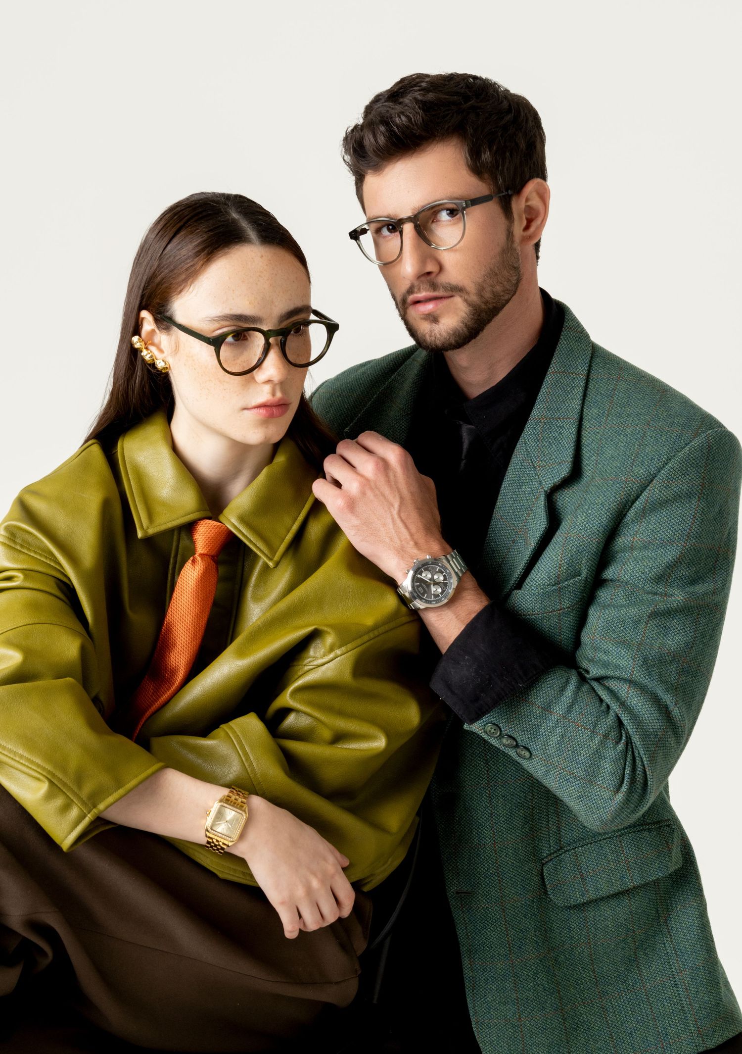 Woman in round glasses and olive jacket sits beside a bearded man in a green blazer, both showing stylish wristwatches.