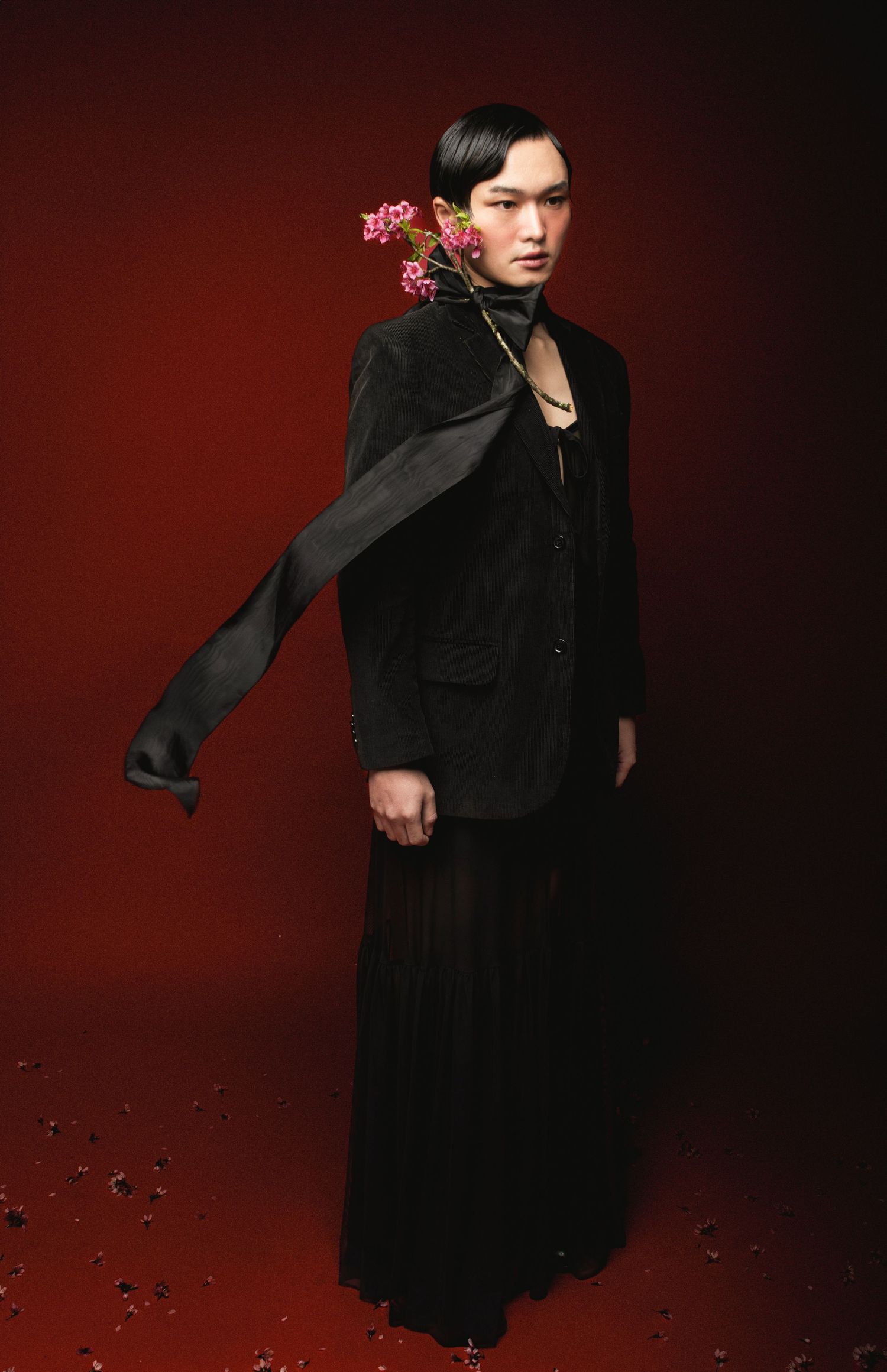 Model in a black velvet jacket and long pleated skirt with a flowing scarf and pink blossoms in the hair against a deep red backdrop.