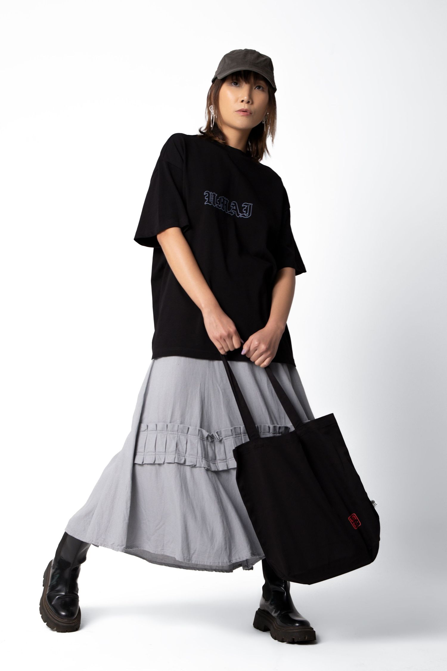 Woman in a black graphic tee, gray tiered maxi skirt, dark cap and lug-sole boots holds a black tote bag with a small red logo.