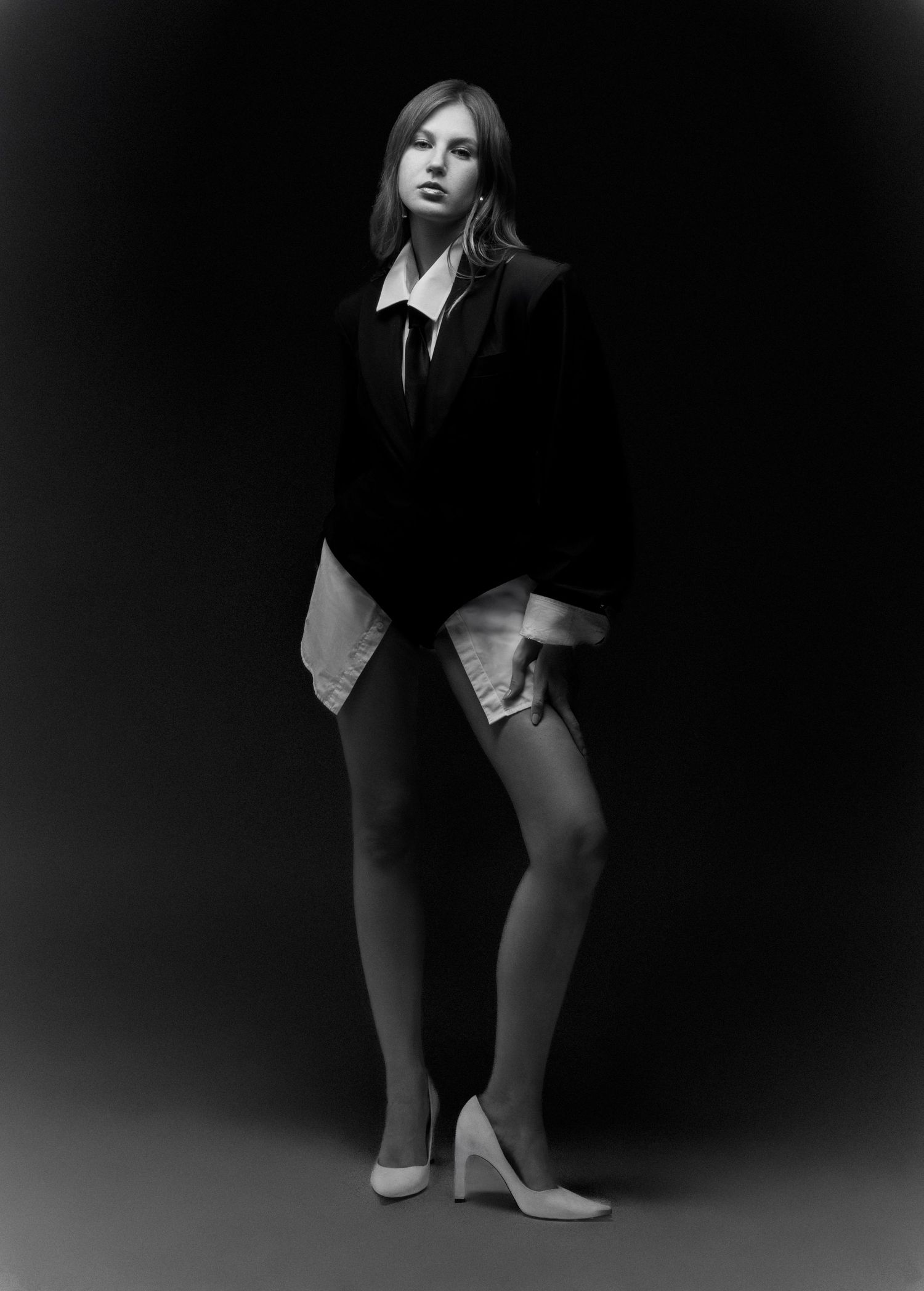 Black and white studio portrait of a model in an oversized black blazer, white shirt, sheer tights and pointed white heels.