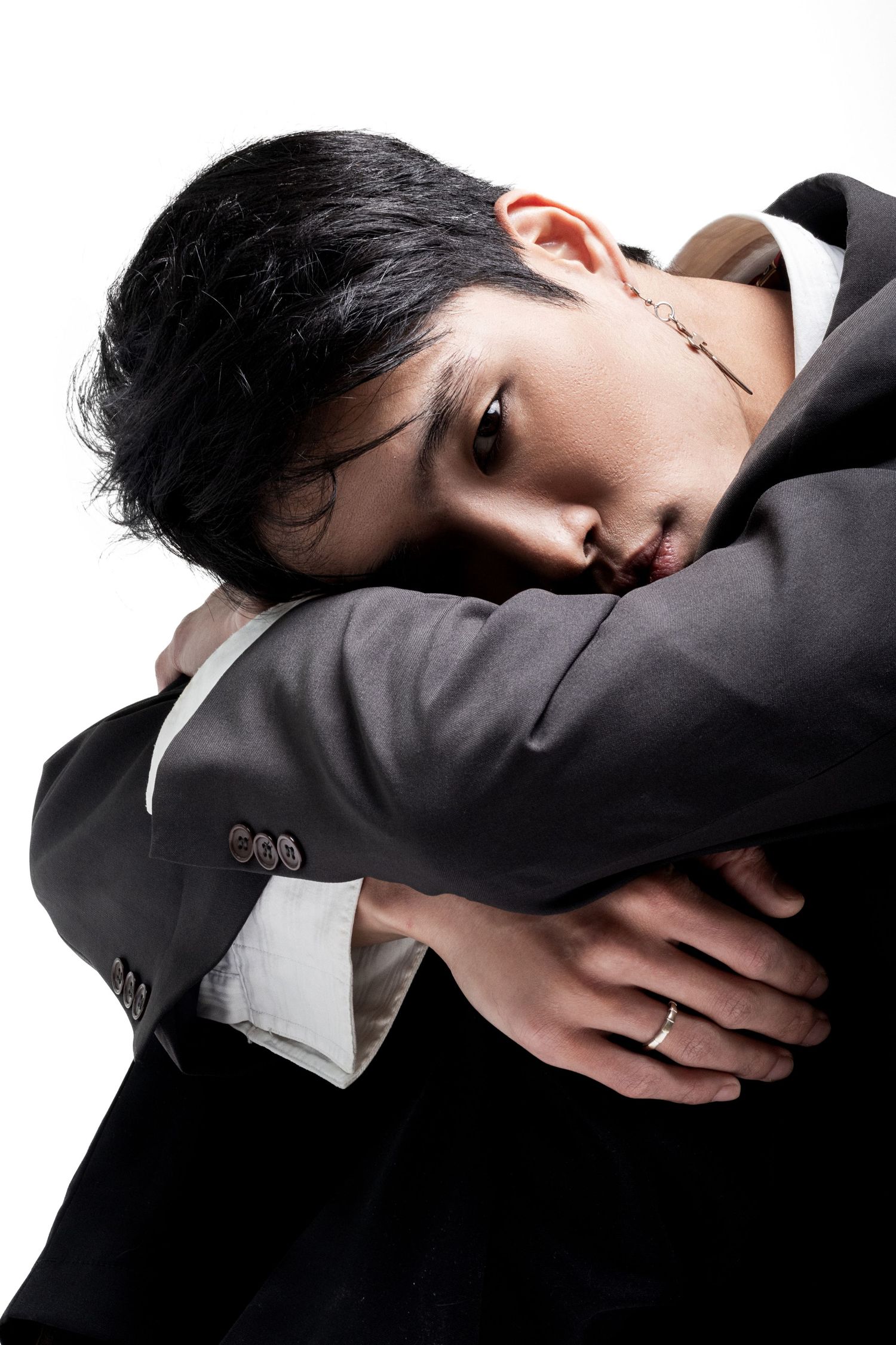 Man in a gray suit rests his head on crossed arms, gazing sideways at the camera, wearing a ring and drop earring.