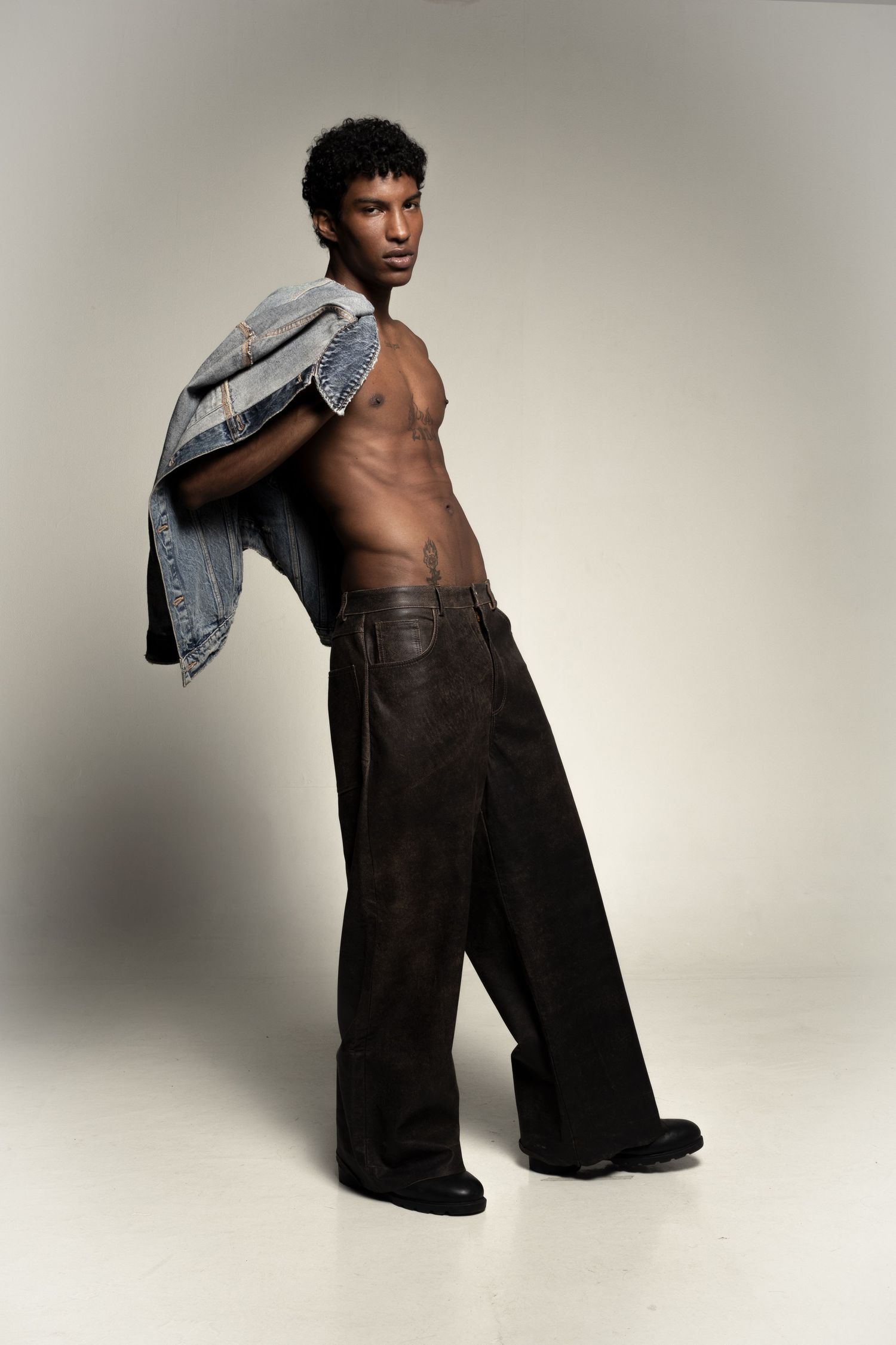 Shirtless young man holds a denim jacket over his shoulder, wearing baggy brown leather pants and black shoes in a studio.
