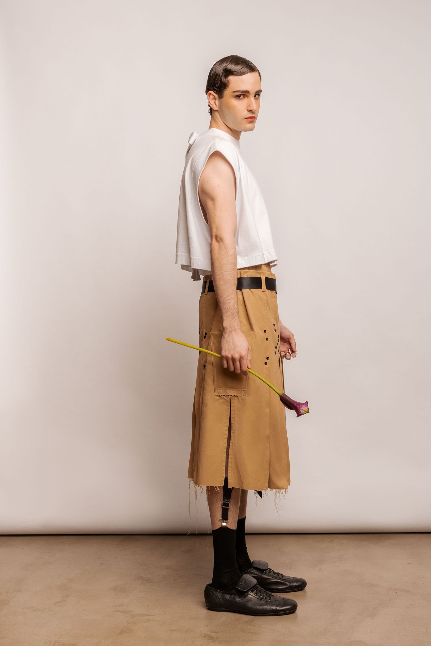 Side view of a model in a white sleeveless top, tan pleated utility kilt and black socks holding a long-stemmed calla lily.