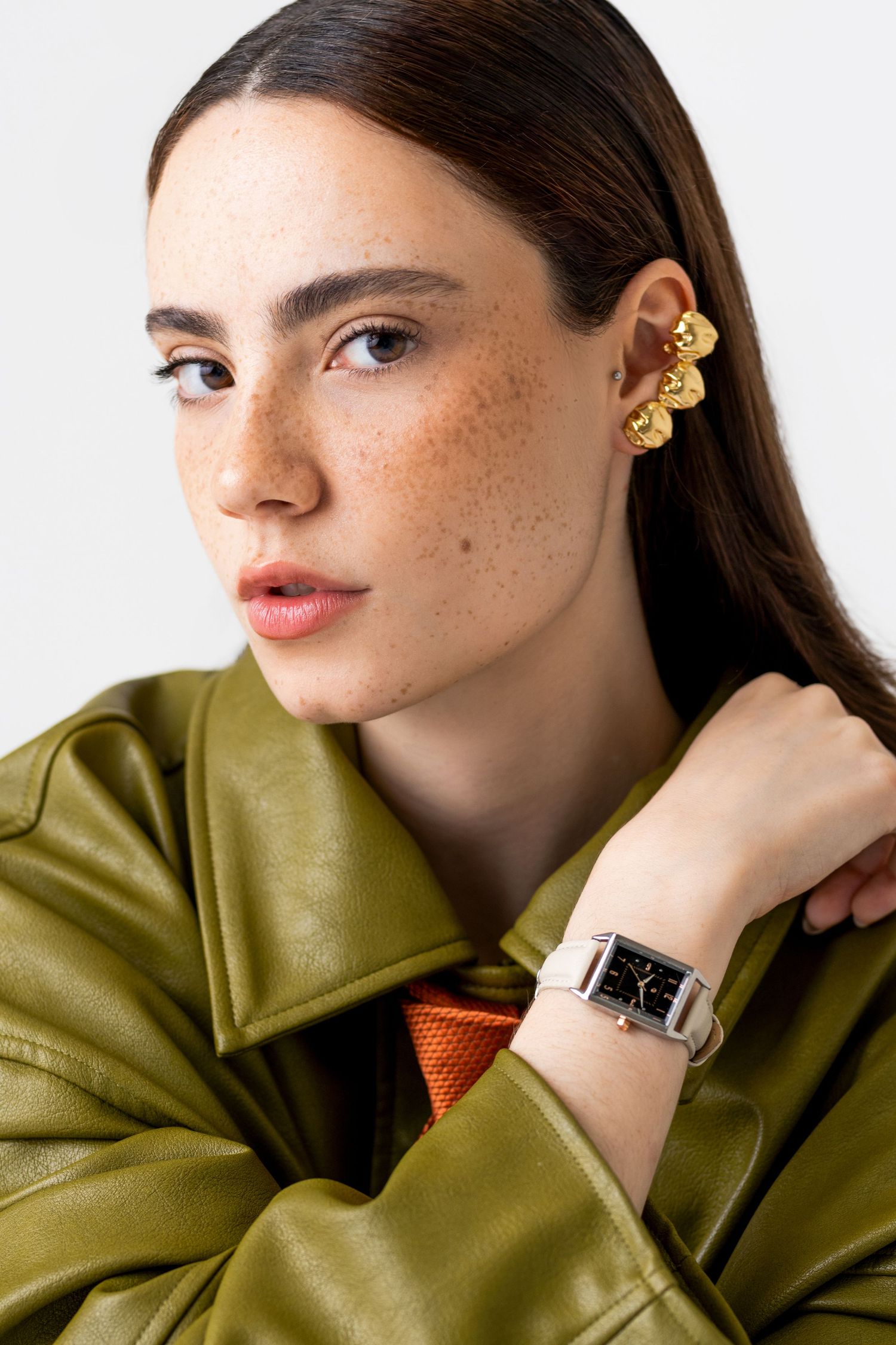 Freckled woman in an olive green leather jacket wears a gold ear cuff and a square-faced wristwatch with a white strap.
