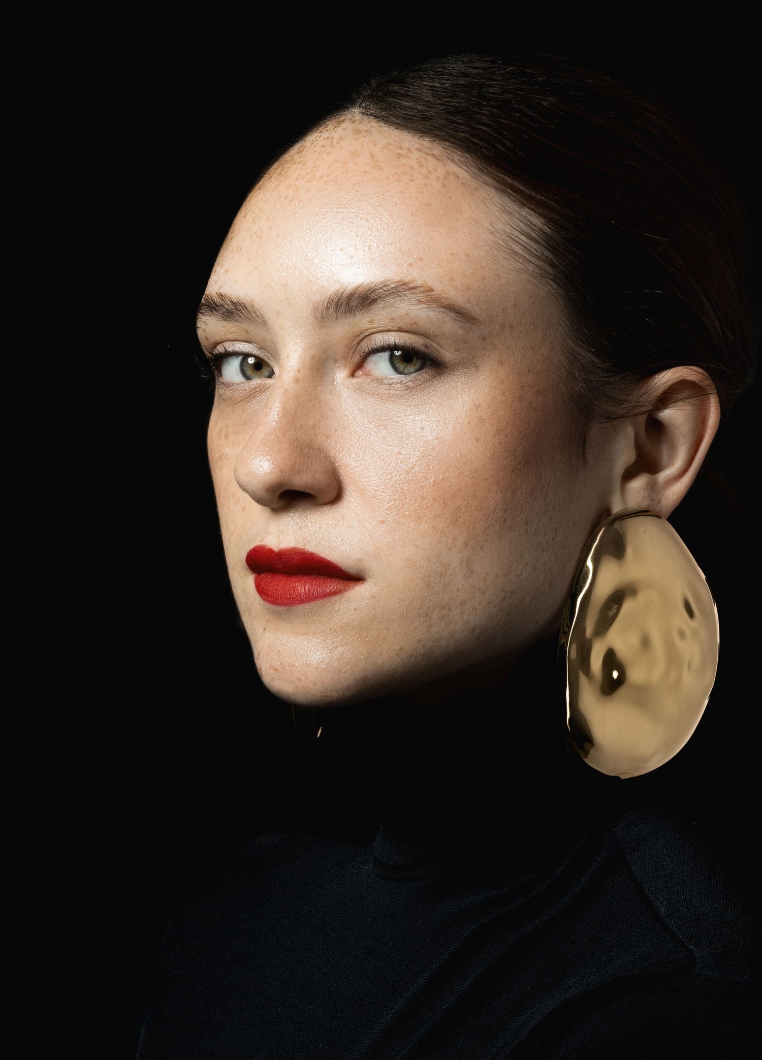 Portrait of a freckled woman with red lipstick, a large hammered gold oval earring and black turtleneck on black.