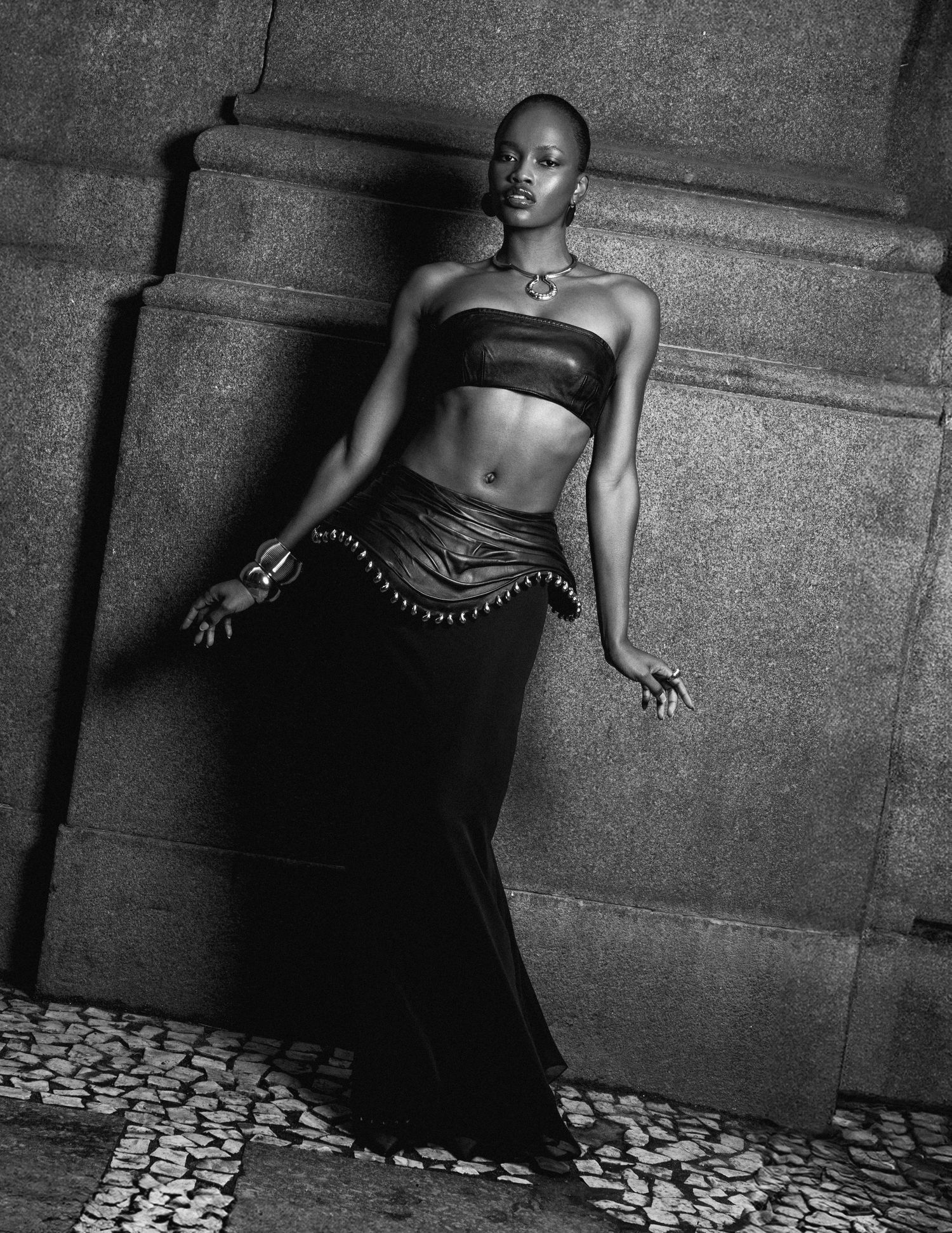 Black and white image of a woman in a leather bandeau top and long skirt leaning against a stone pillar on a cobblestone ground.