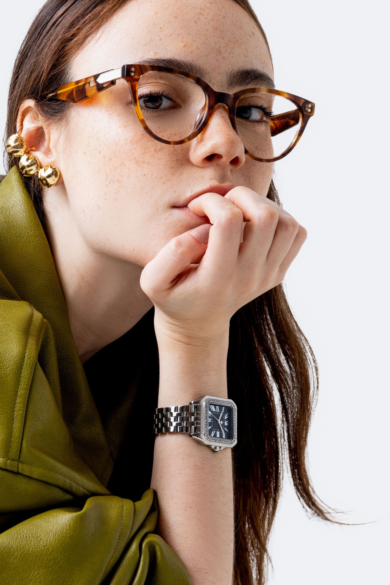 Woman in tortoiseshell glasses and gold ear cuff rests her chin on her fist, wearing a silver rectangular watch with a blue dial.