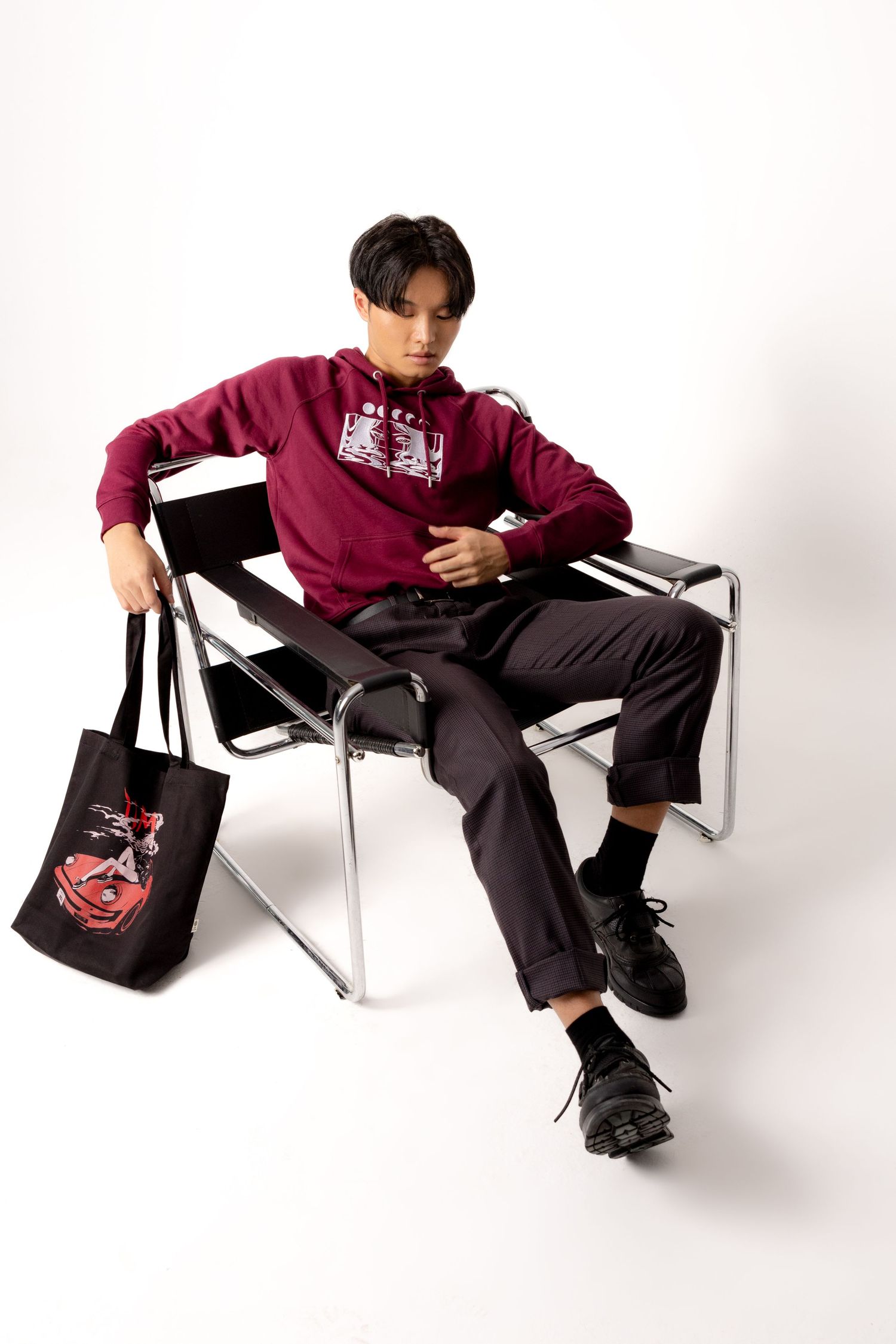Man in a burgundy hoodie, gray cuffed trousers and black shoes lounges in a tubular steel chair beside a black car-print tote bag.