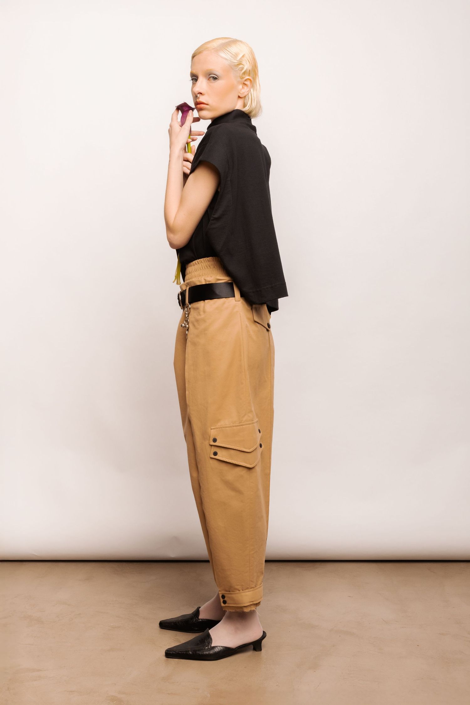 Side view of a blonde model in a black cropped top and tan cargo pants with leg pockets, holding a dark flower and wearing black mules.