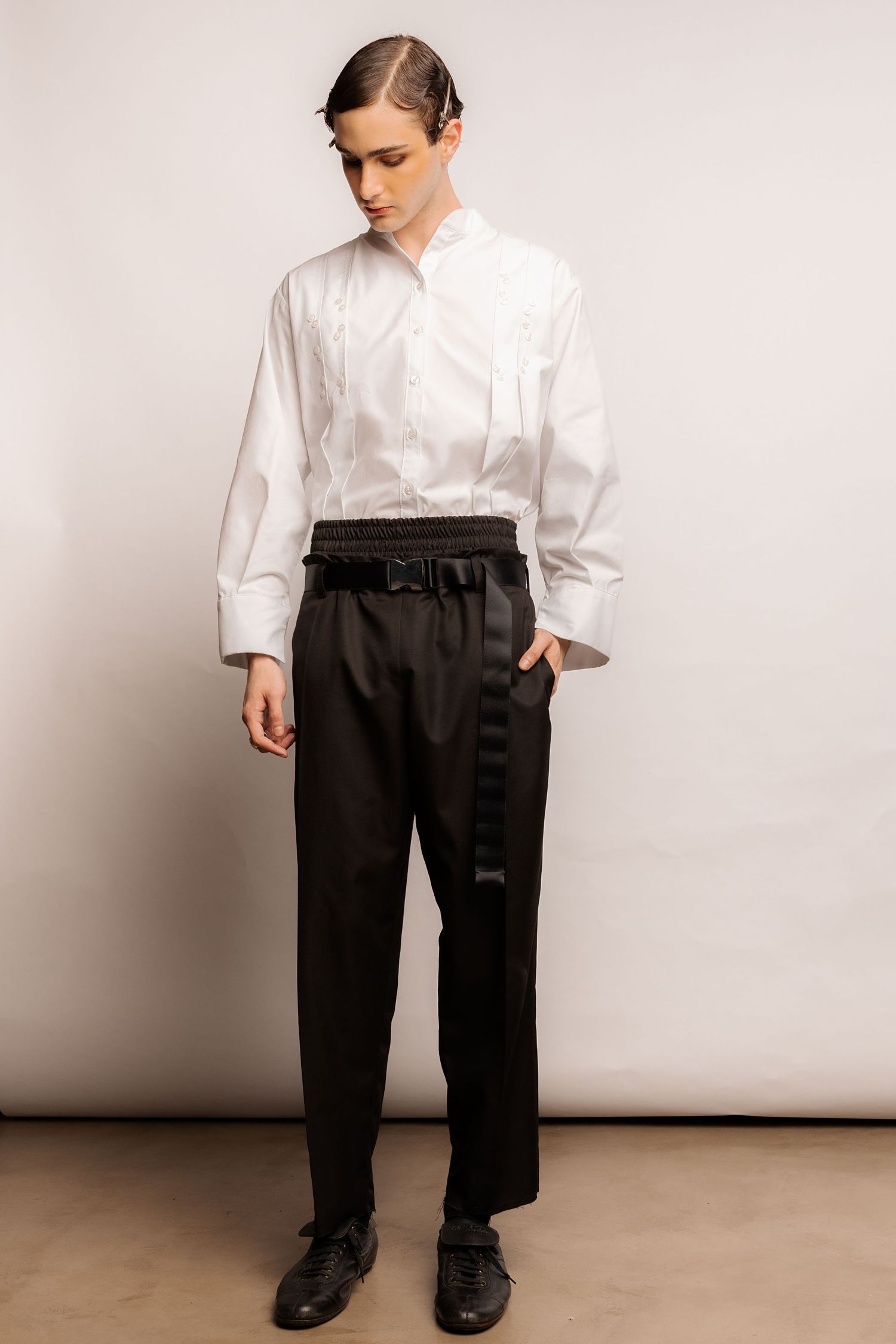 Model wearing a white pleated tuxedo-style shirt with black high-waisted trousers, a utility buckle belt and black lace-up shoes.