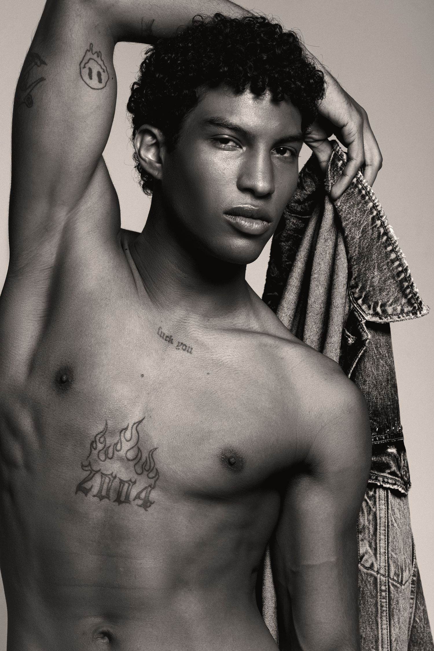Shirtless model with curly hair raises one arm while holding a denim jacket, showing a flame tattoo on his chest in black and white.