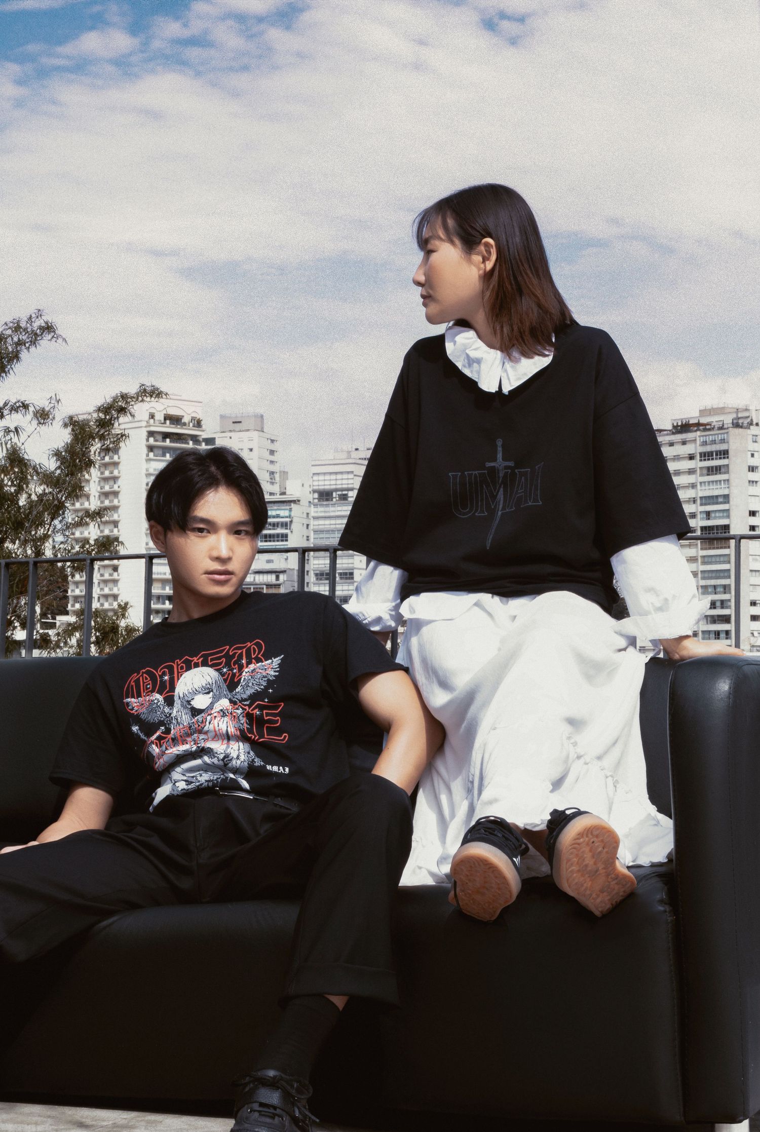 Two young people in black graphic streetwear sit on a black leather sofa on a sunny rooftop with city buildings behind them.