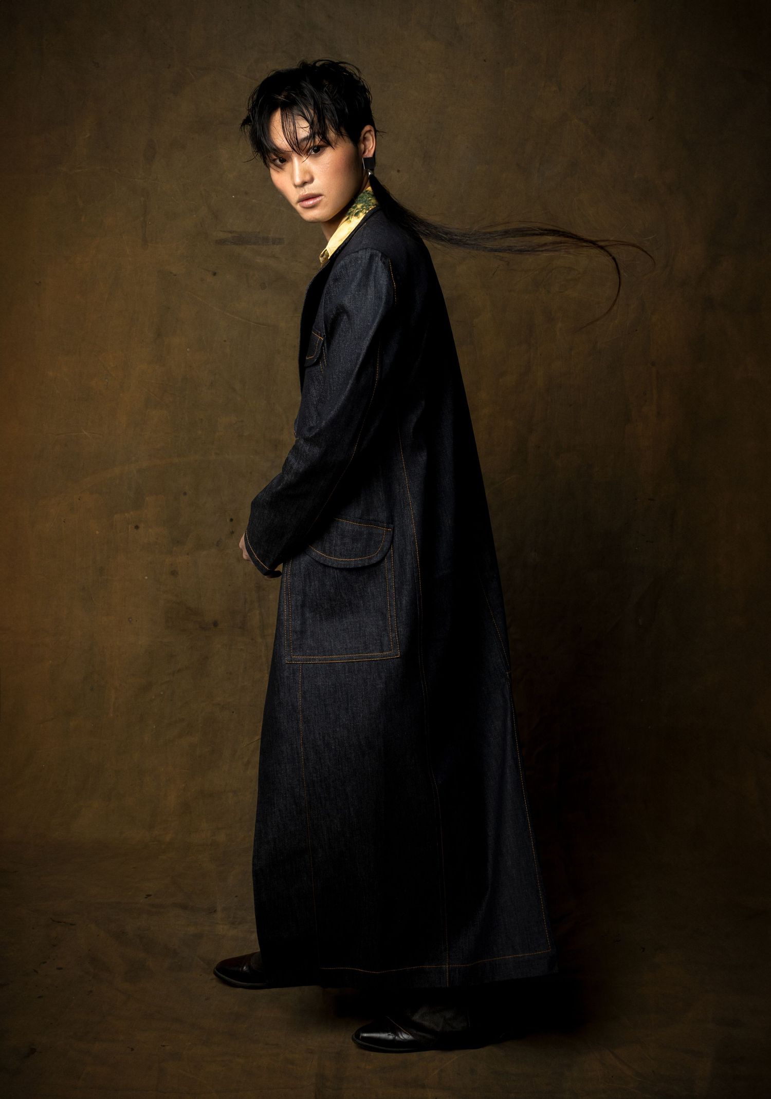 Model in a long dark denim coat with hair flowing sideways stands in profile against a moody golden-brown studio backdrop.