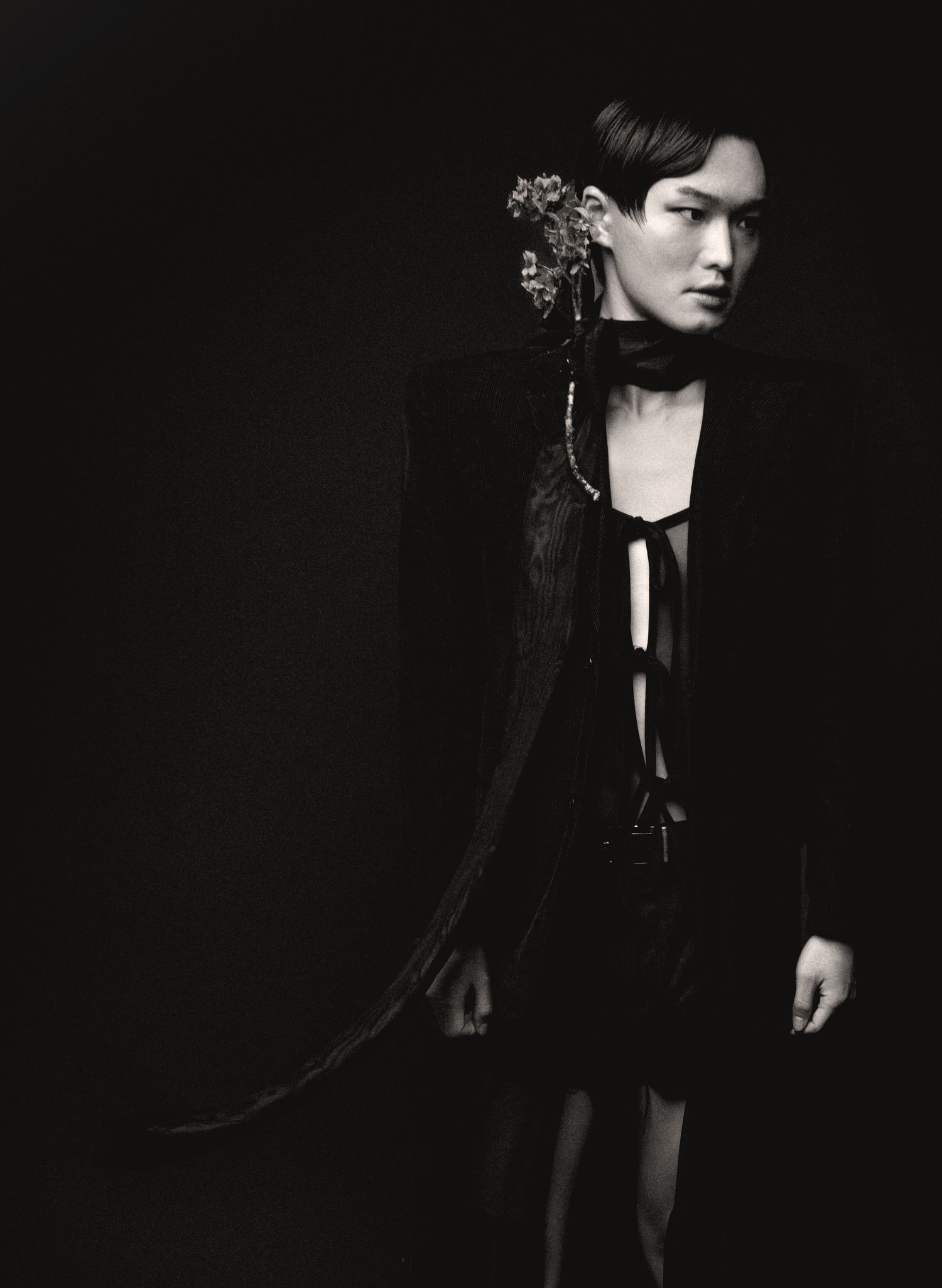 Dramatic black and white portrait of a model in a dark tie-front top and jacket with a floral ornament in the hair against a black backdrop.
