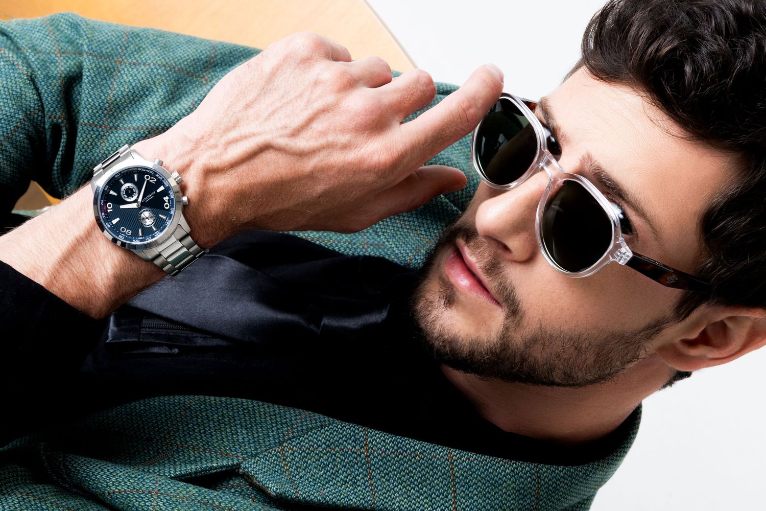 Man in a green tweed jacket and round sunglasses adjusts his stainless steel wristwatch with a blue dial.