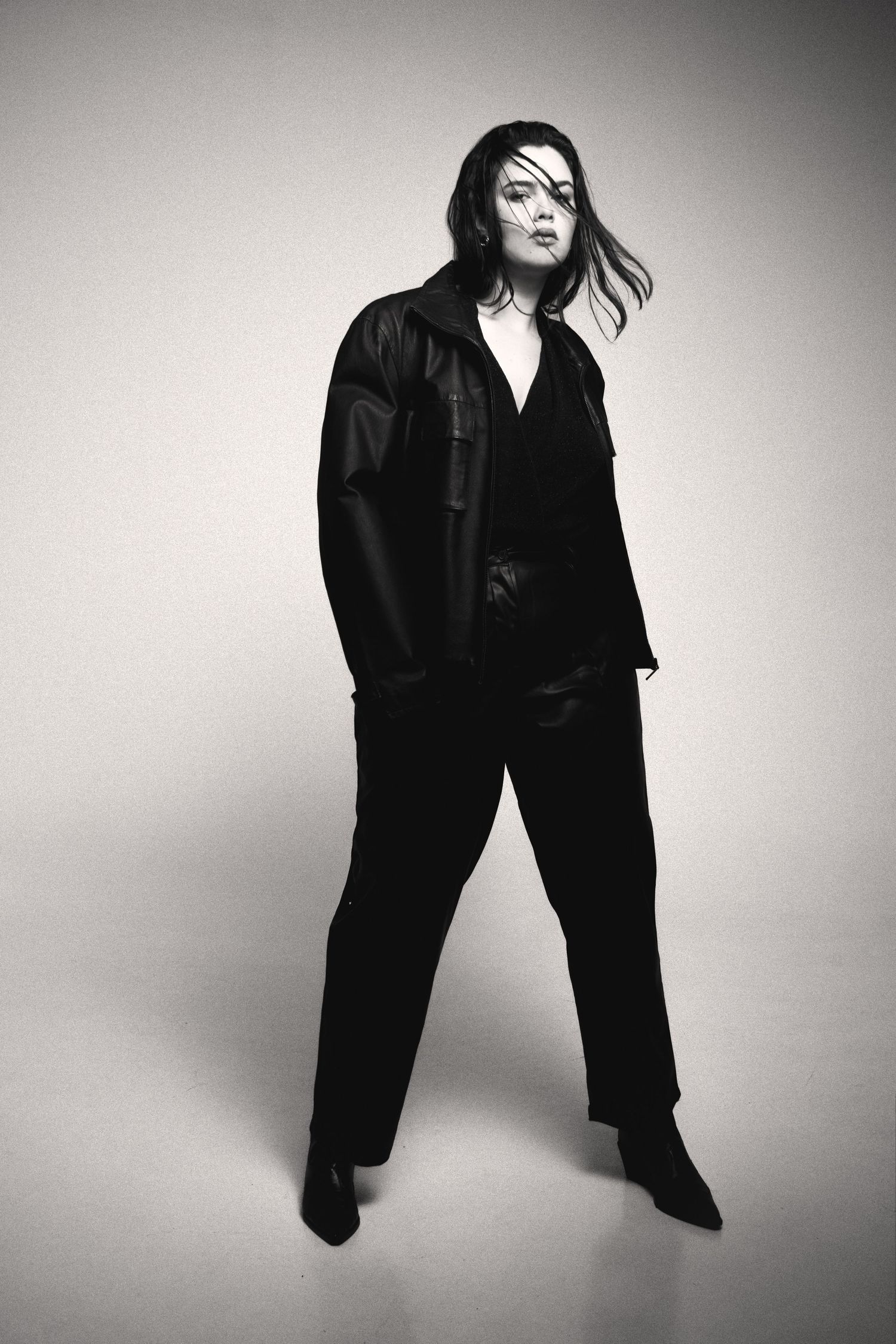 Full-length black and white photo of a model in a black leather jacket and trousers with windswept hair, standing in a wide stance.
