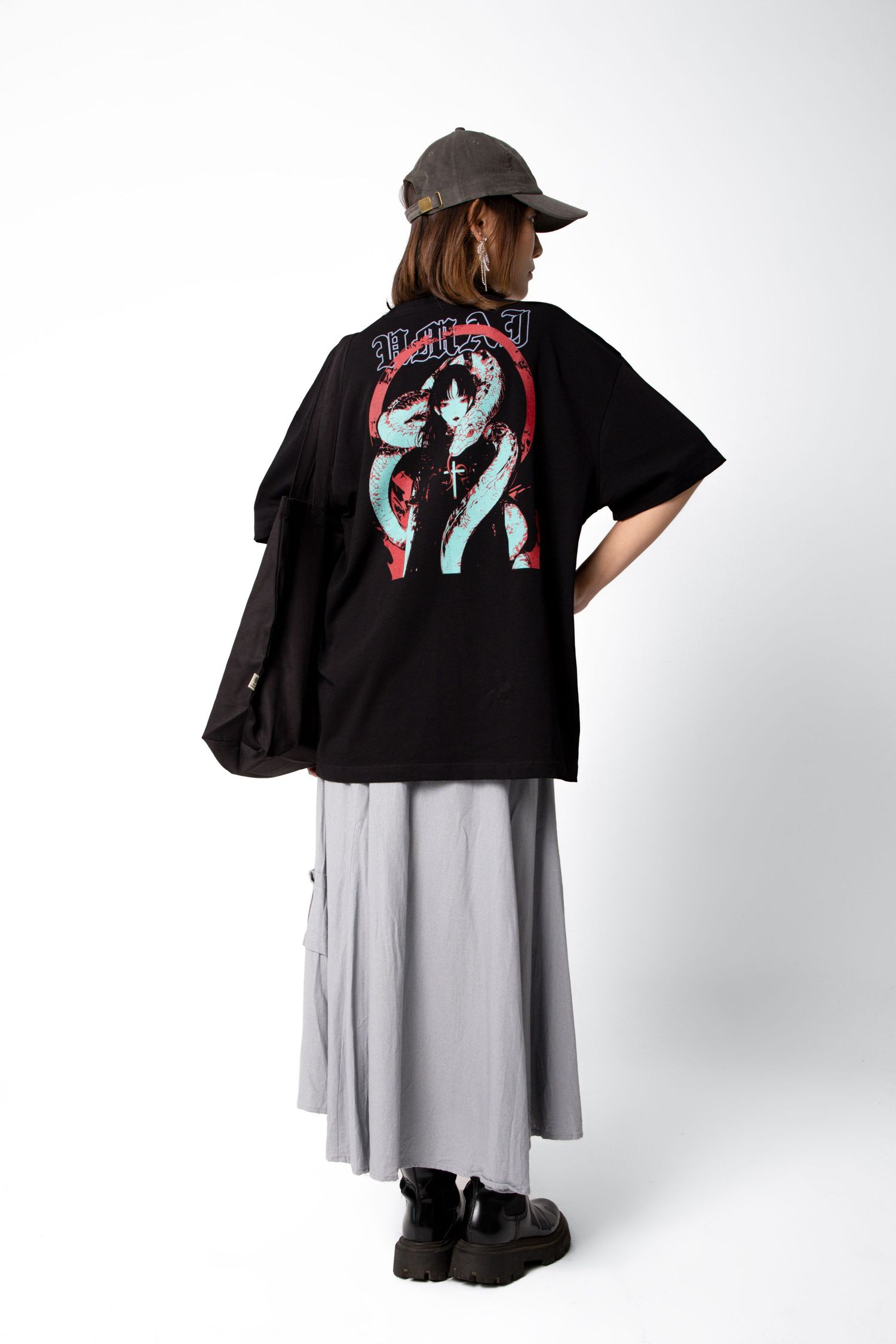 Back view of a model in a black oversized graphic tee with a red and teal print, gray cargo skirt, olive cap and chunky black boots.