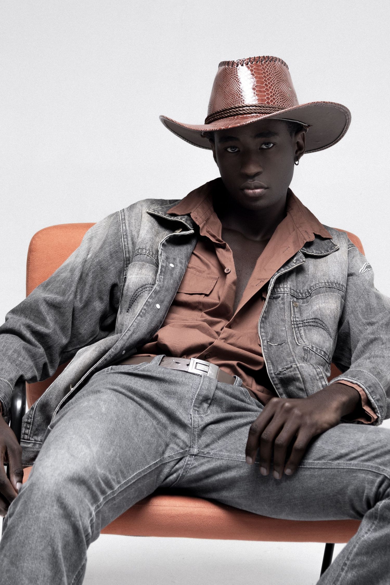 Male model lounges in an orange chair wearing a brown cowboy hat, rust button-up shirt, gray denim jacket and matching jeans with a belt.