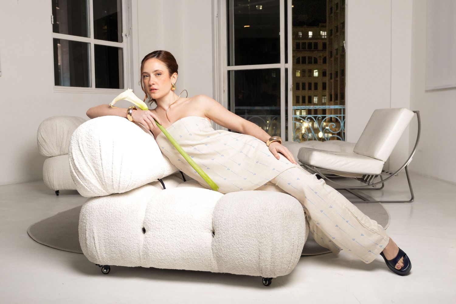 Model reclines on a plush white sofa in a cream strapless top and matching pants, holding a green calla lily in a bright loft.