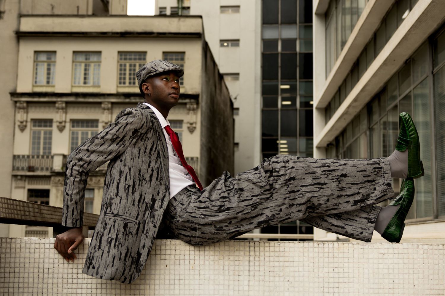 Man in a patterned suit, flat cap and green shoes reclines on a ledge with city buildings rising behind him in an urban fashion shoot.