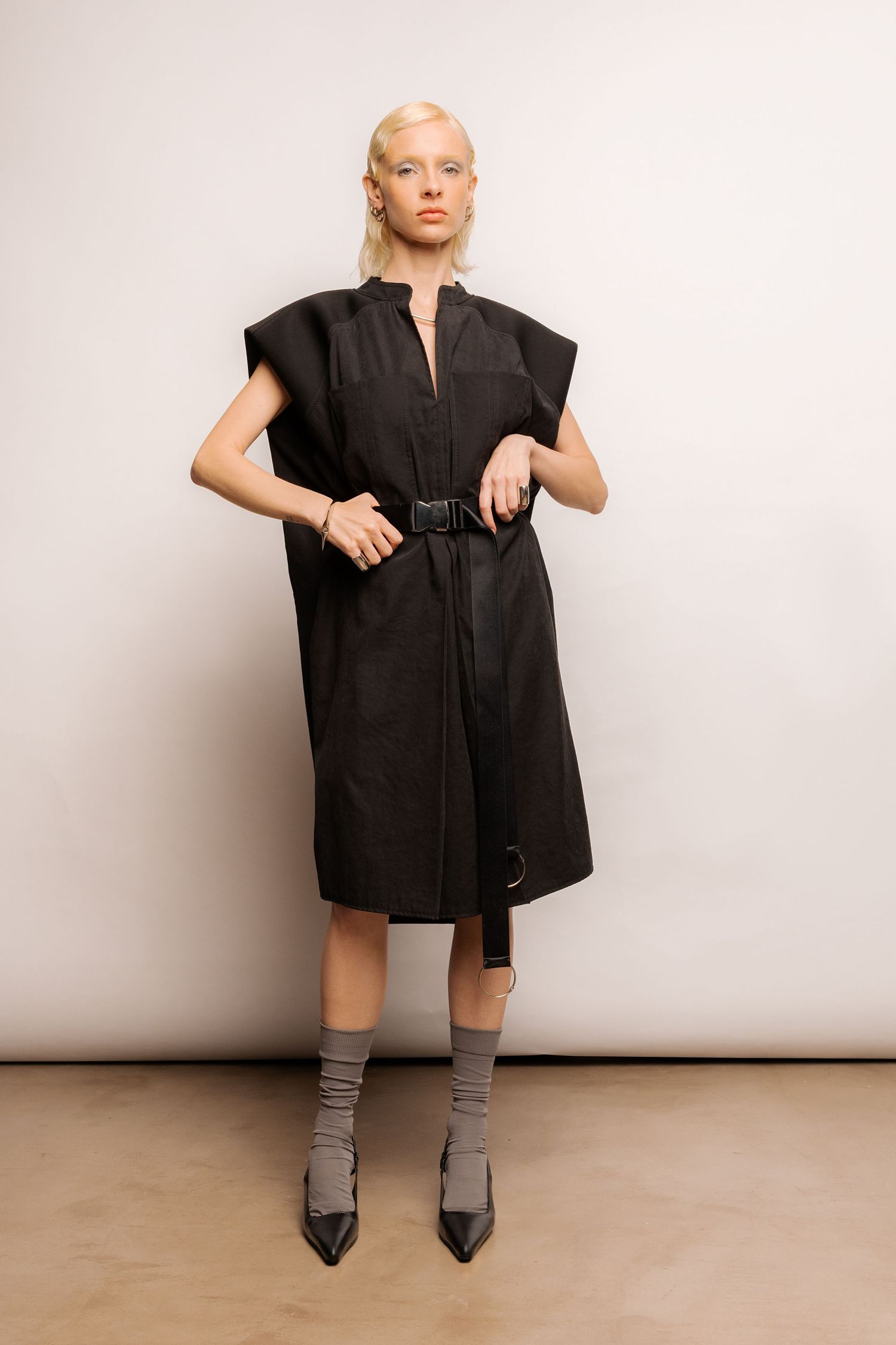 Model in a black sleeveless oversized belted dress with structured shoulders, grey socks and pointed black heels.