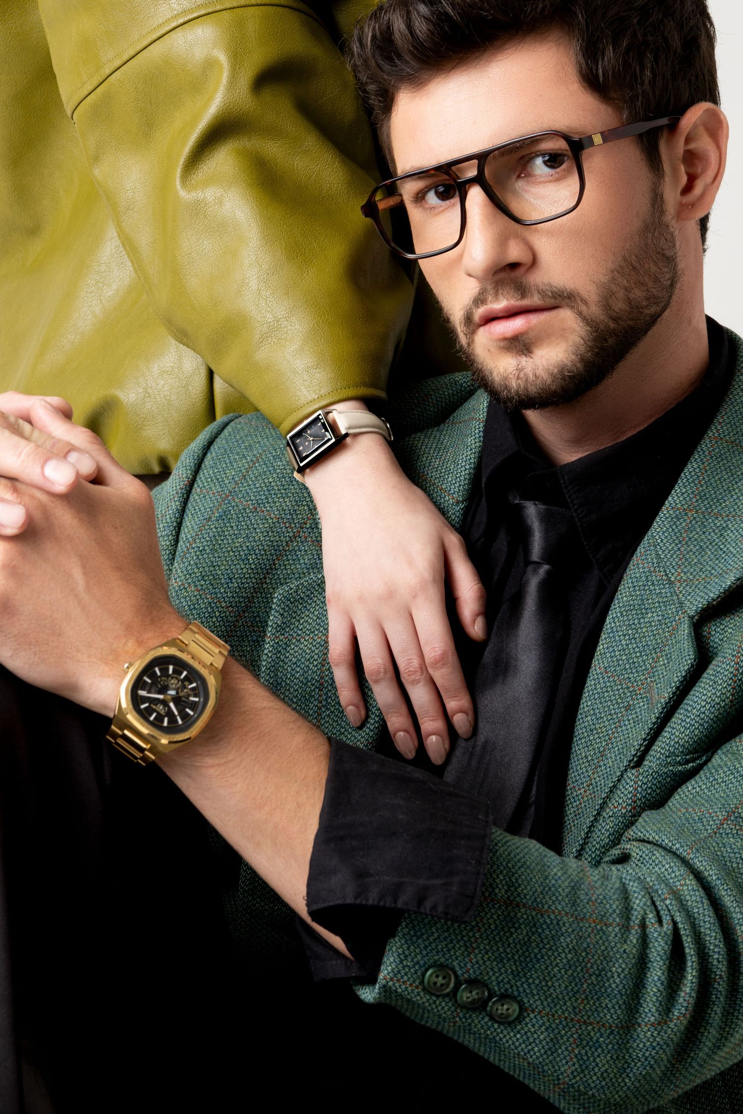 Bearded man in glasses and a green tweed blazer crosses his arms, showing a gold watch while a second watch is visible nearby.