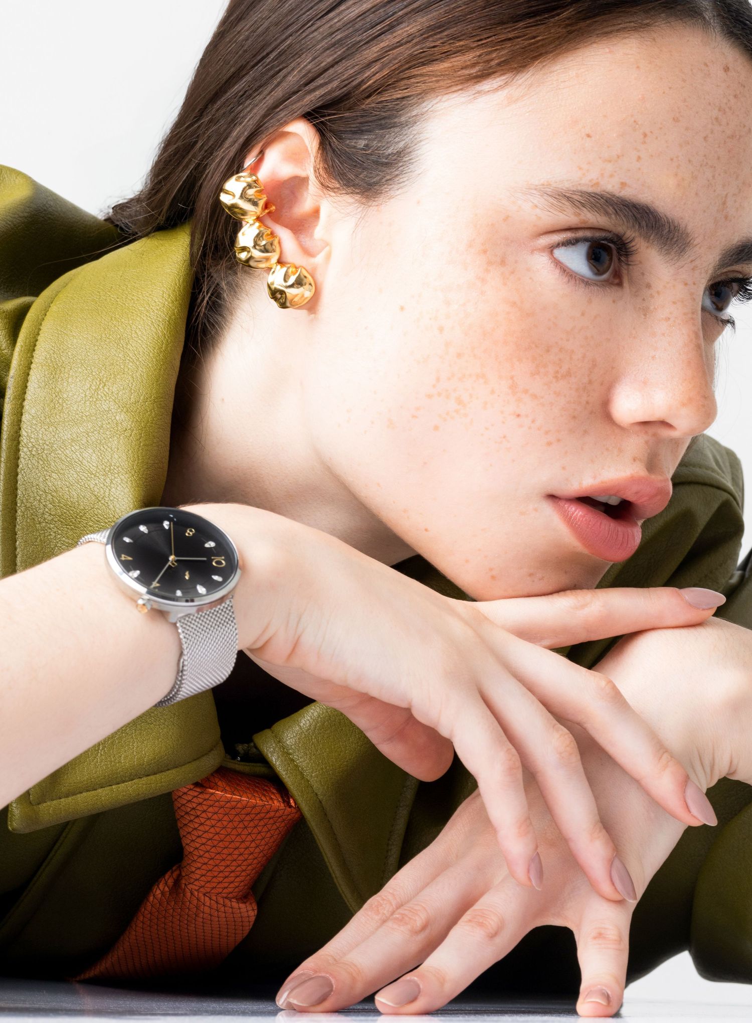 Woman in an olive leather jacket and gold ear cuff leans on her arms, showing a black-dial watch with a silver mesh strap.