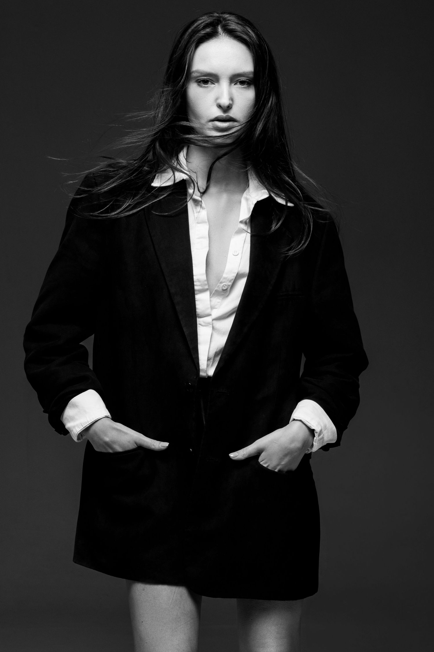 Black and white portrait of a model with windswept hair in an oversized black blazer over a white shirt, hands in pockets.