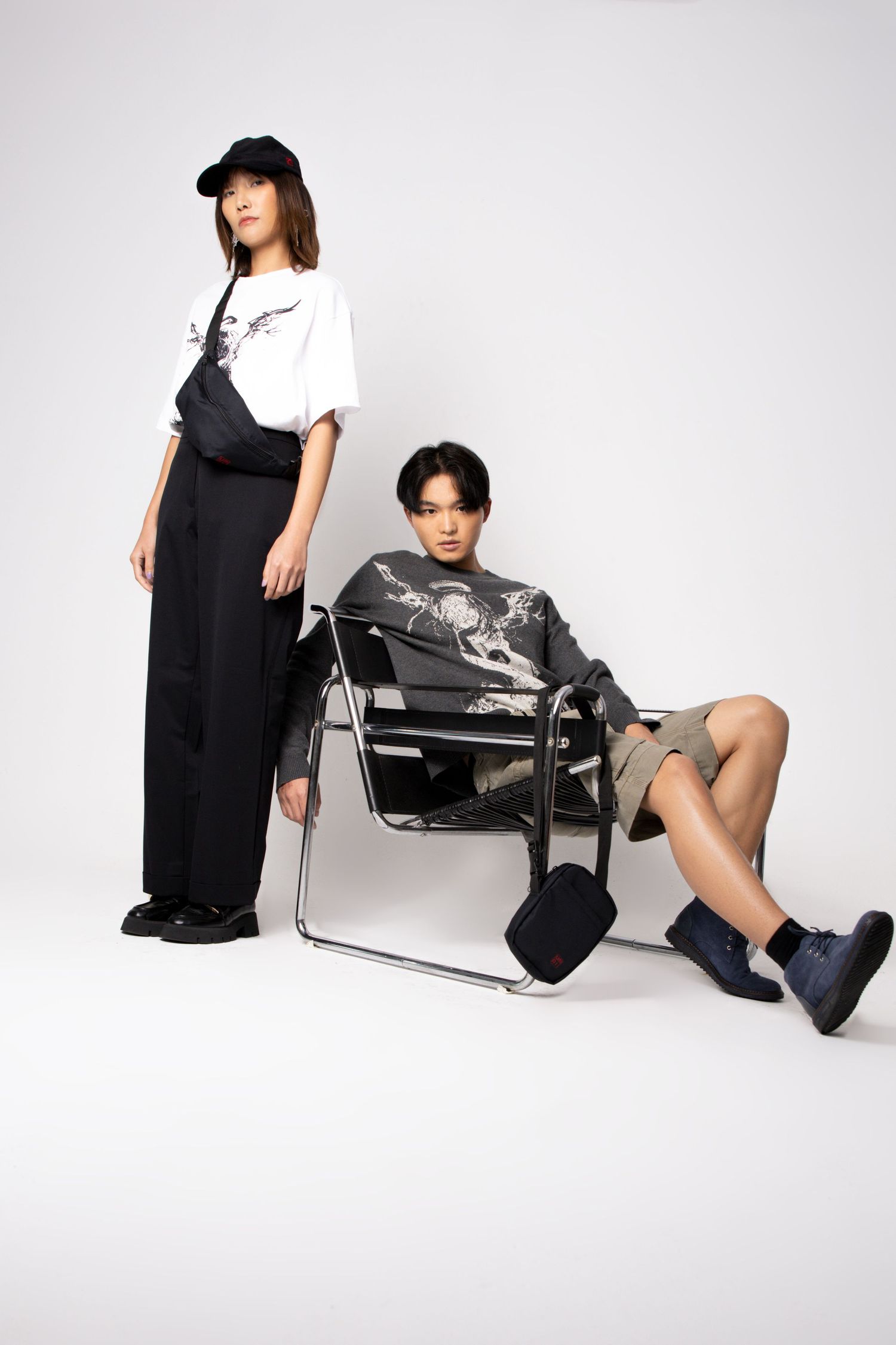 A model in a black cap and wide-leg trousers stands beside a seated model on a chrome chair in graphic streetwear.