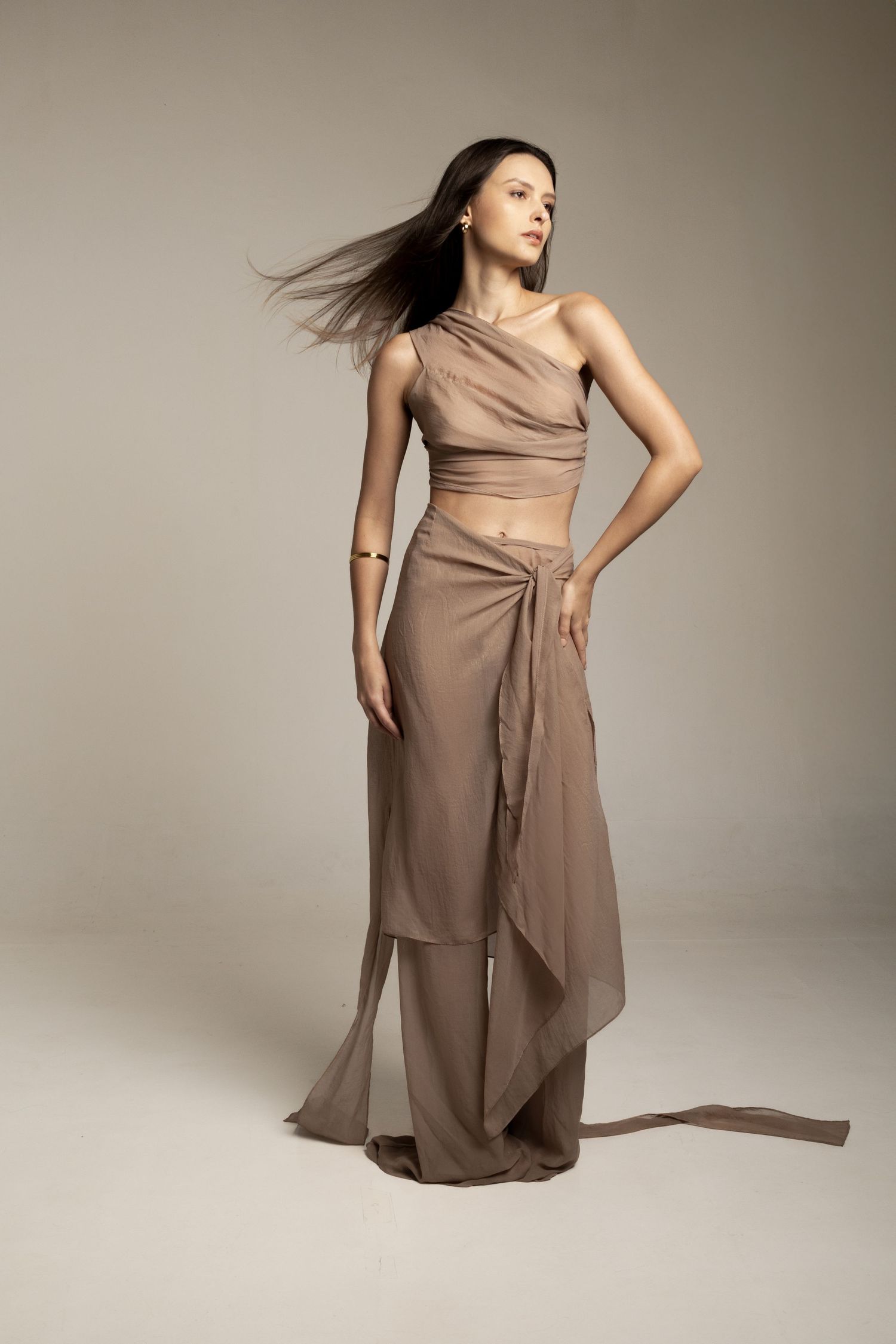 Model in a taupe one-shoulder crop top and draped wrap skirt with wide-leg trousers, hair blowing in a studio.