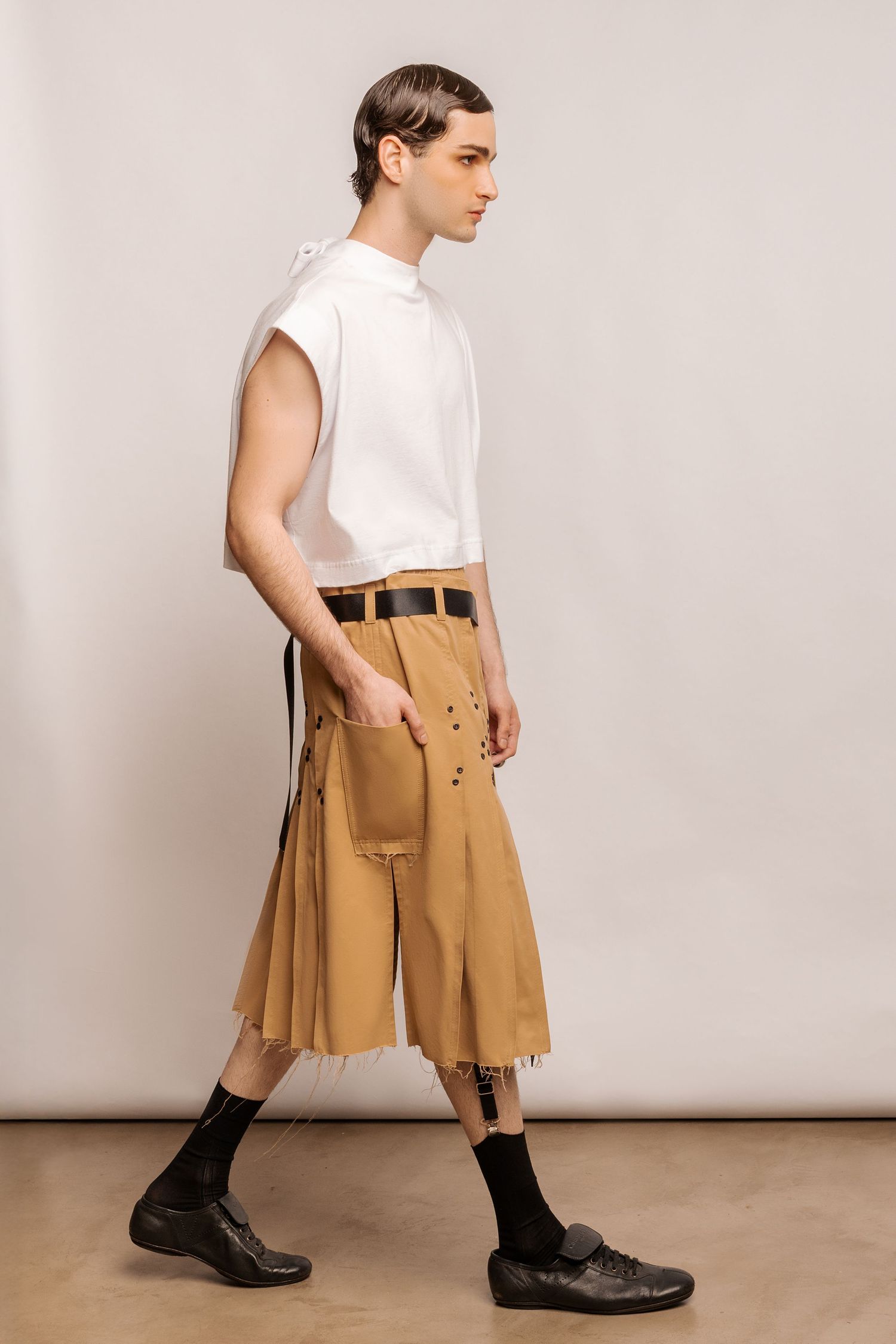 Model in profile wearing a white cap-sleeve top, tan pleated kilt with grommet details and front pocket, black socks and leather shoes.