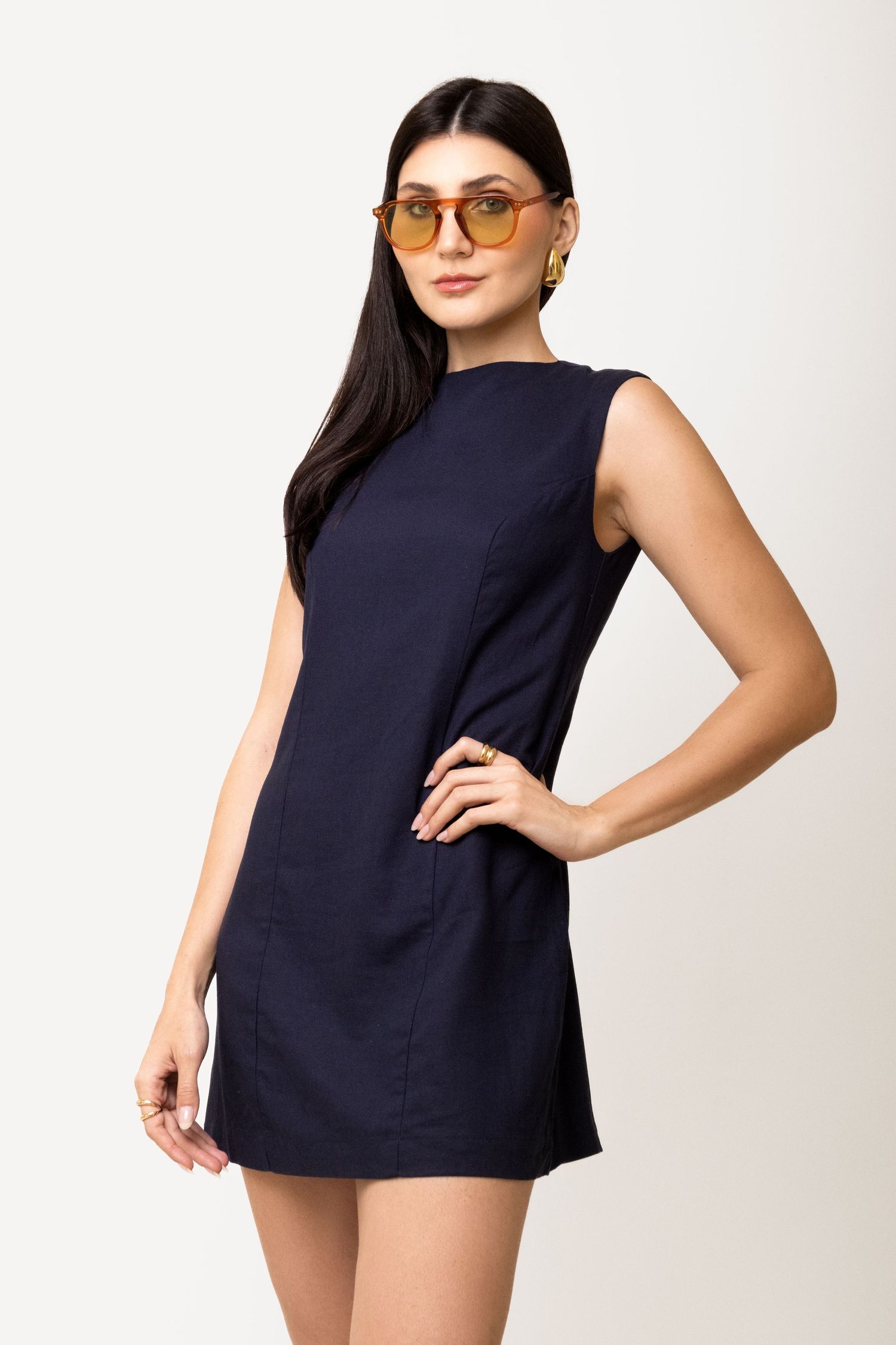 Woman in a navy sleeveless mini shift dress and orange tinted sunglasses poses with one hand on her hip.