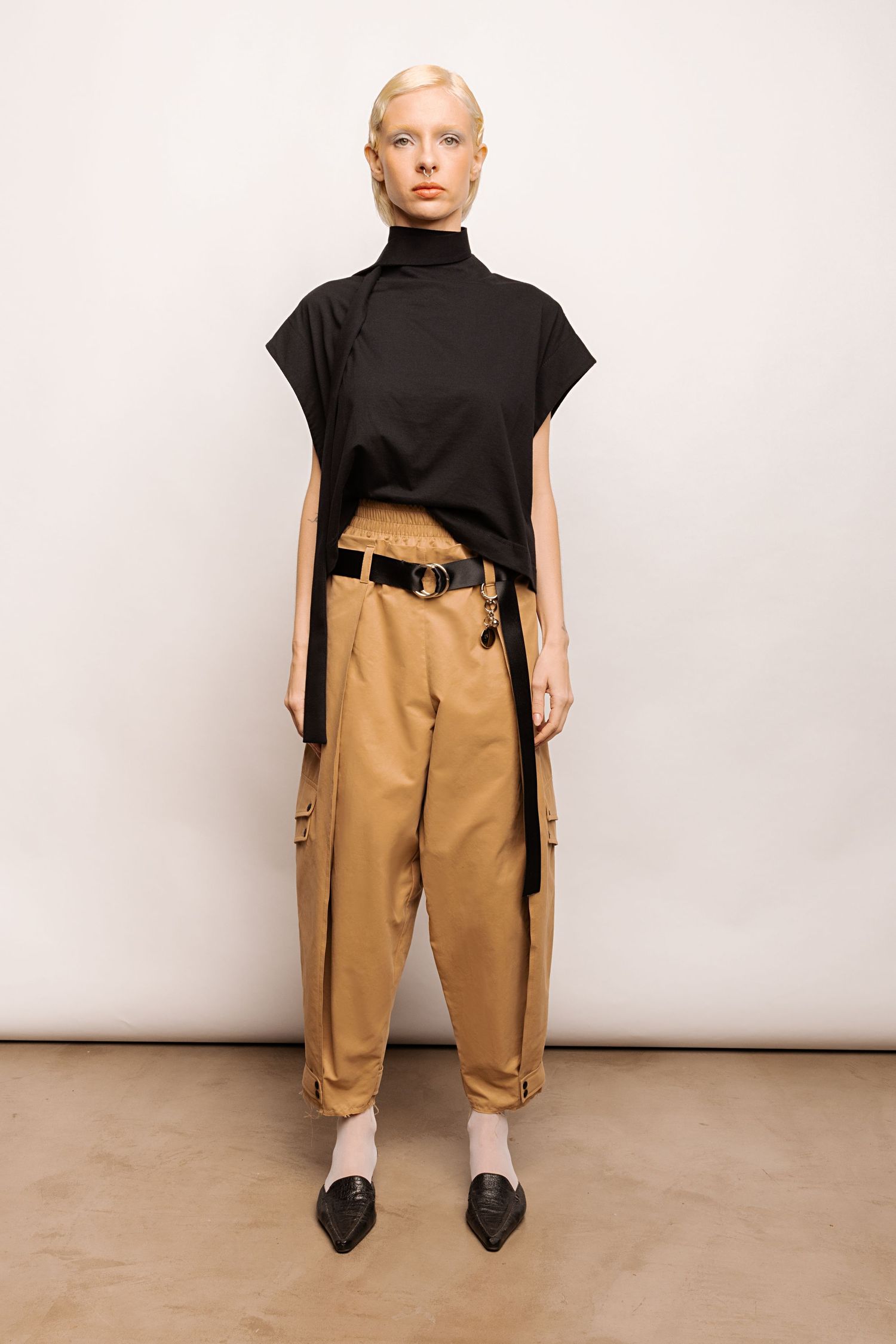 Model wearing a black sleeveless funnel-neck top with tan cargo trousers, a black belt and pointed black flats against a plain backdrop.