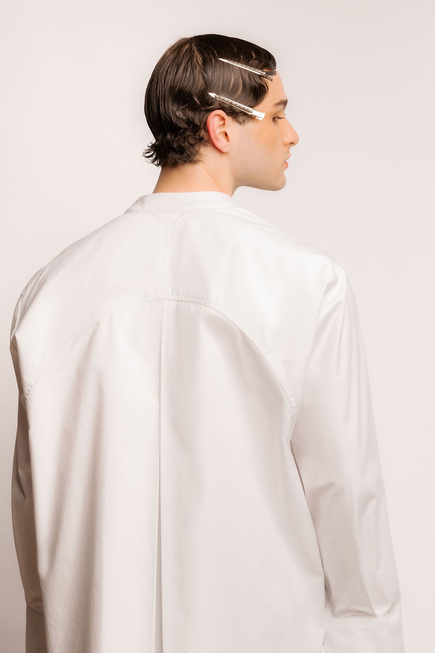 Back view of a model in an oversized white shirt with a yoke seam and decorative metallic hair clips in slicked-back hair.