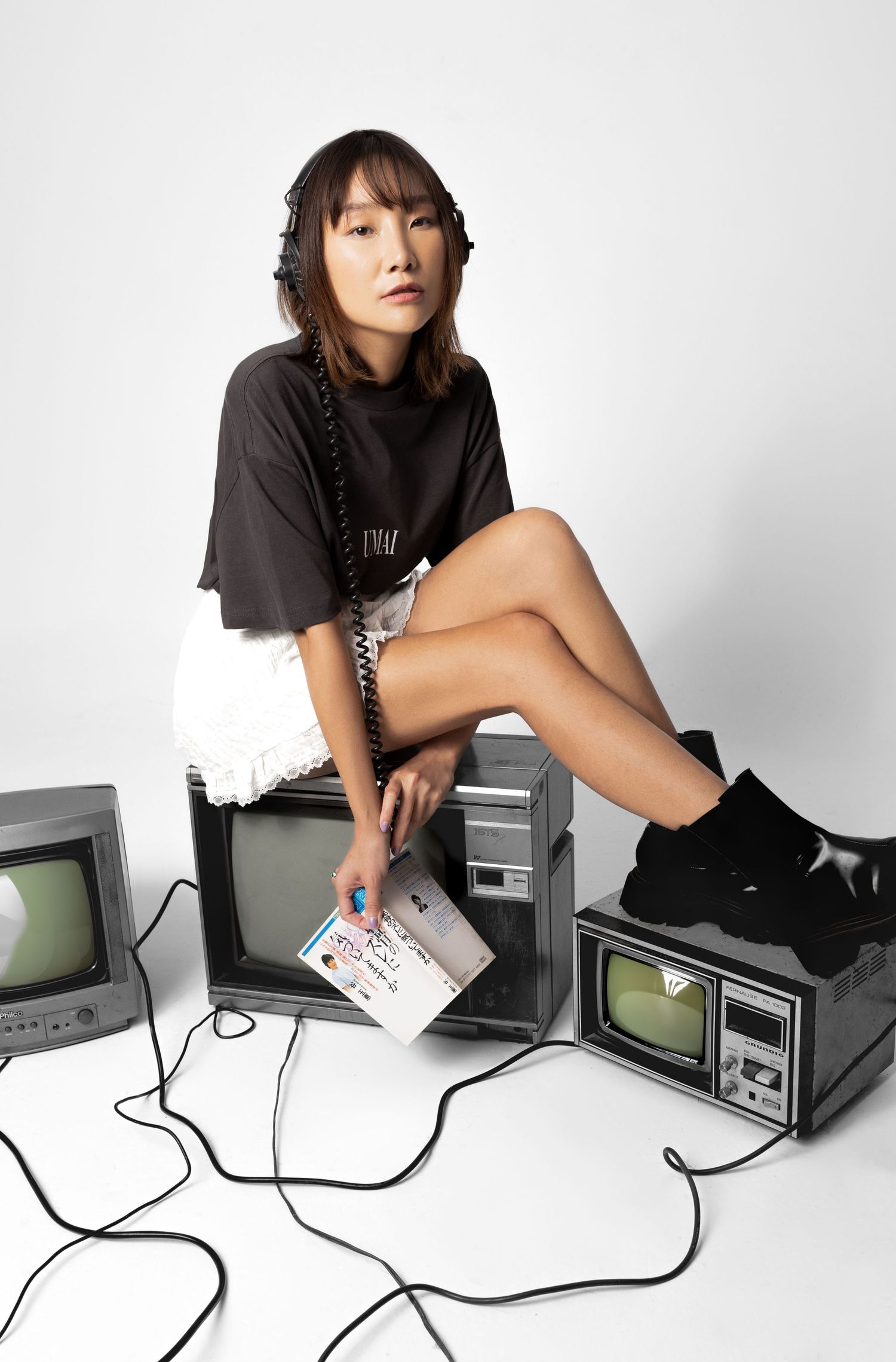 Woman with a braid, headphones and black socks sits on retro TVs with tangled cables, holding a colorful booklet in a studio.