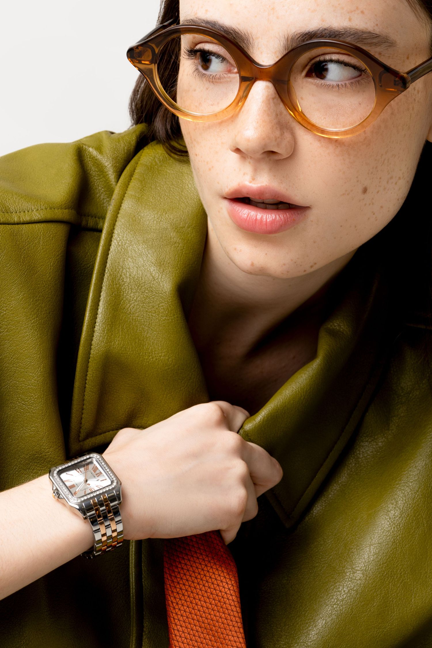 Woman in round amber glasses and an olive leather jacket glances aside, wearing a two-tone rectangular watch on her wrist.