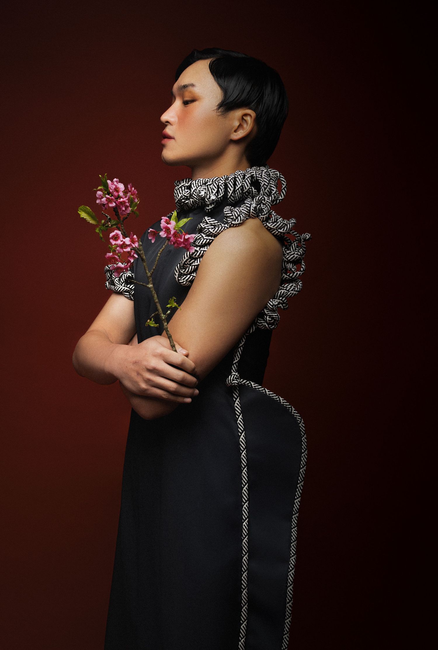 Woman with short dark hair in a black dress with ruffled braided trim holds pink blossoms against a deep red background.