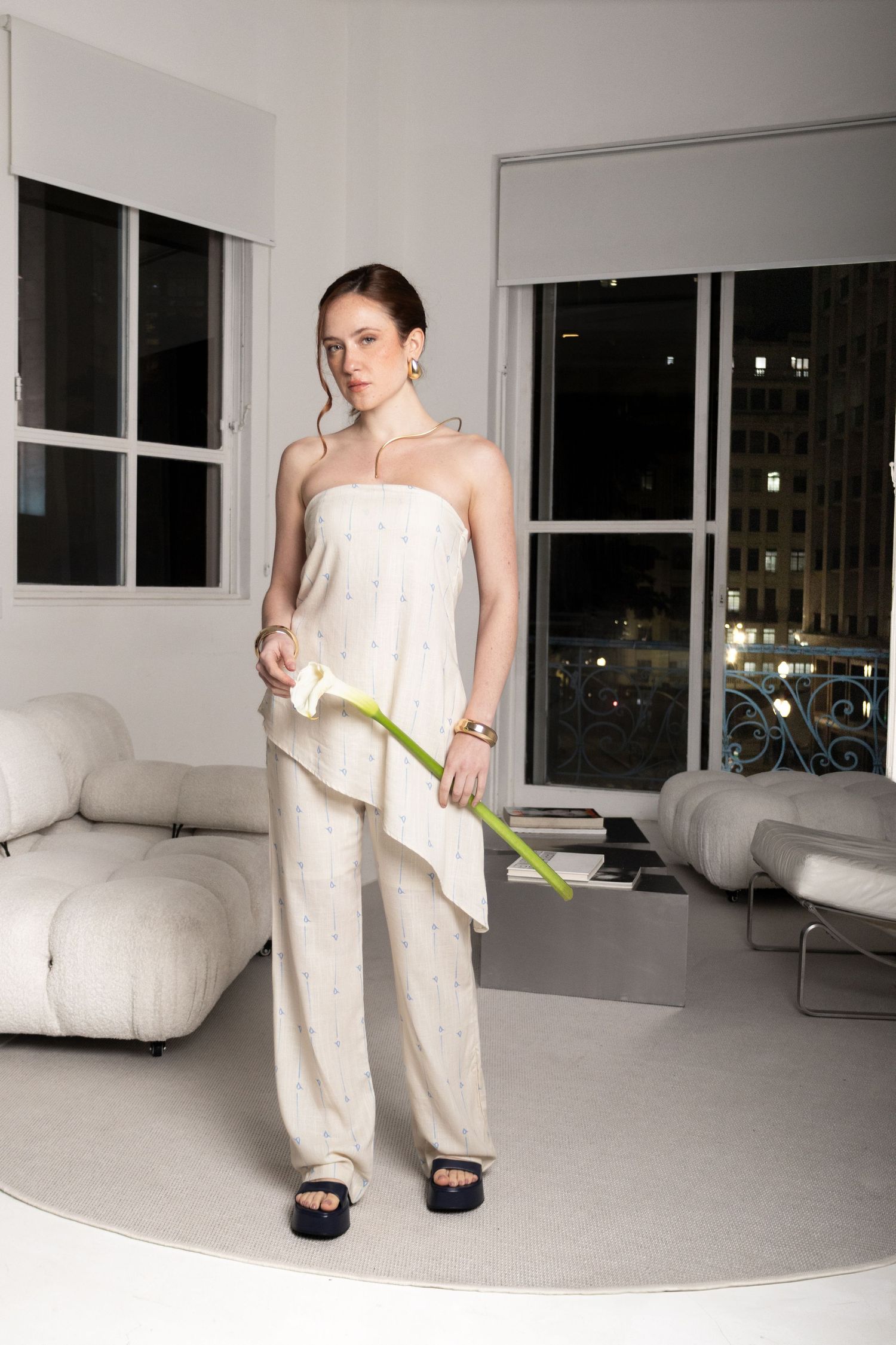 Woman in a cream strapless top and patterned trousers holds a calla lily in a modern living room with city views at night.