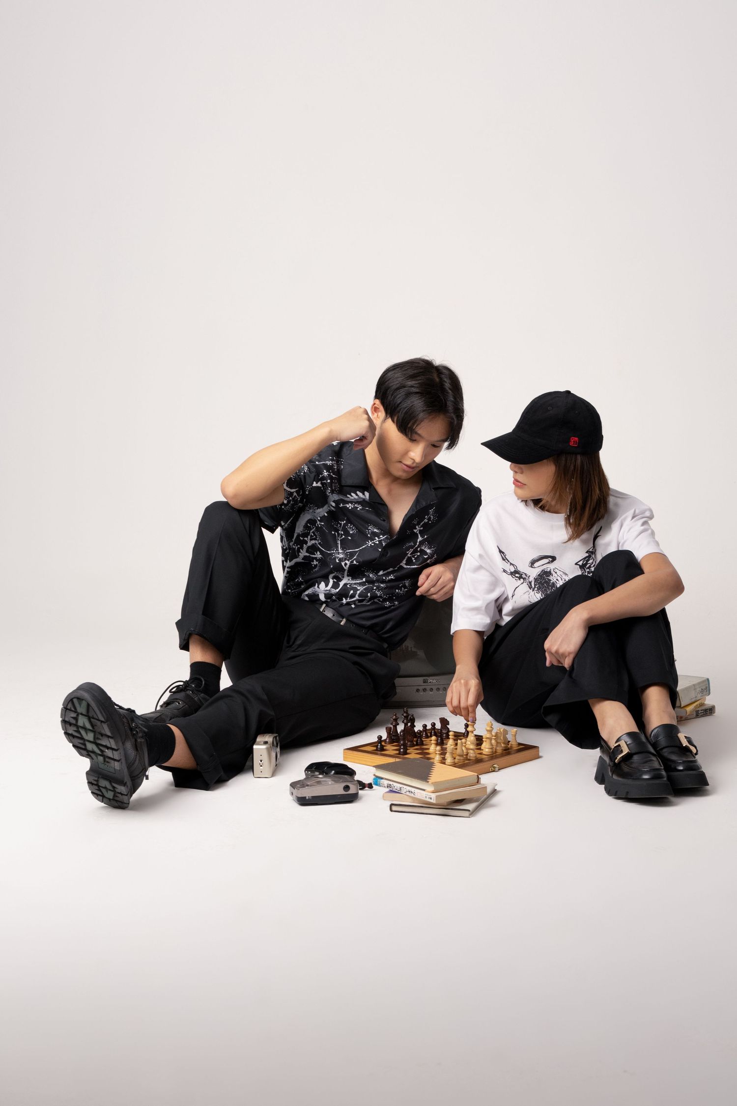 Two models in black streetwear sit on a white studio floor playing chess, with books and a small camera beside them.